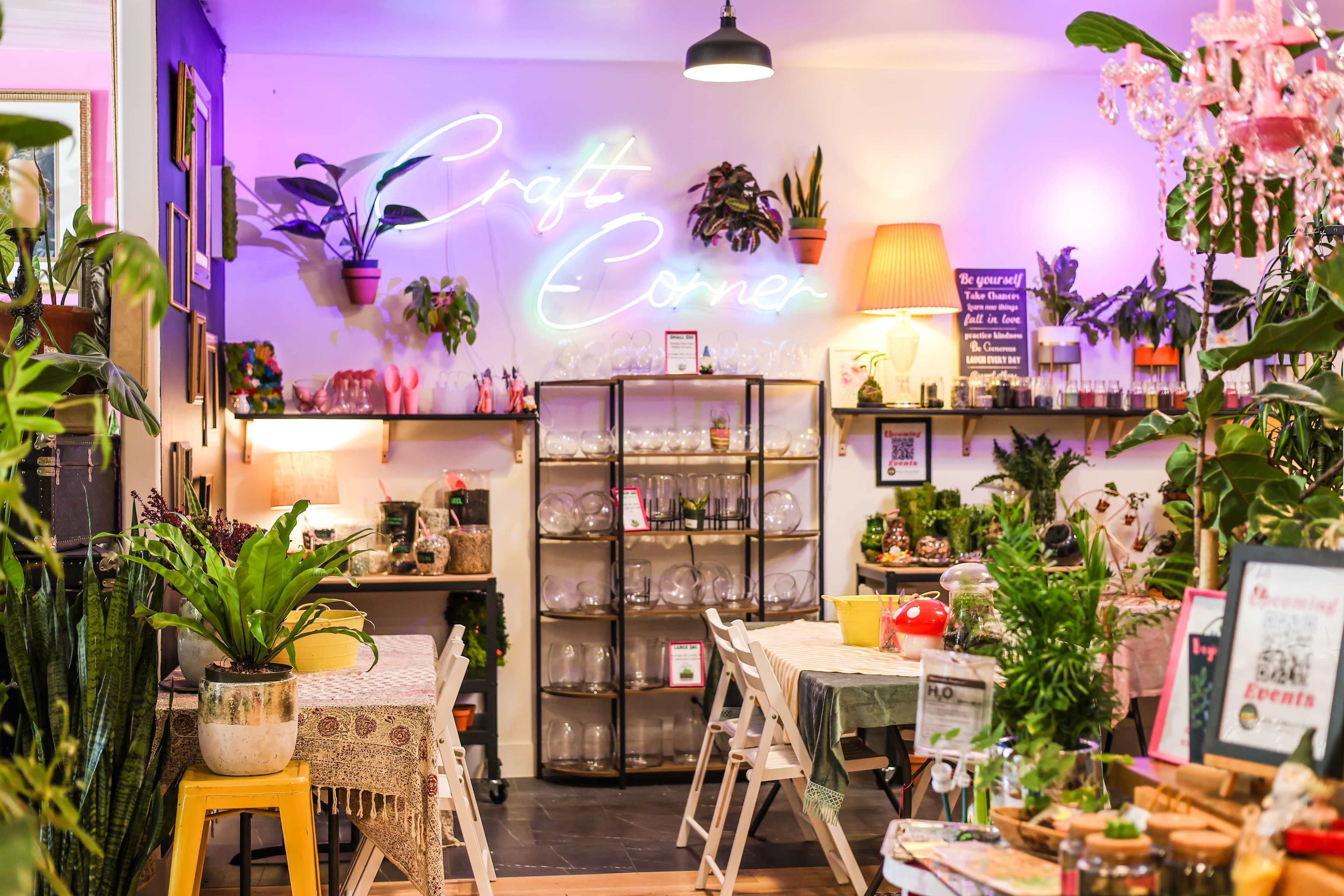 A cozy craft corner filled with plants, tables, and shelves displaying various craft supplies and decorations, illuminated by a neon sign.