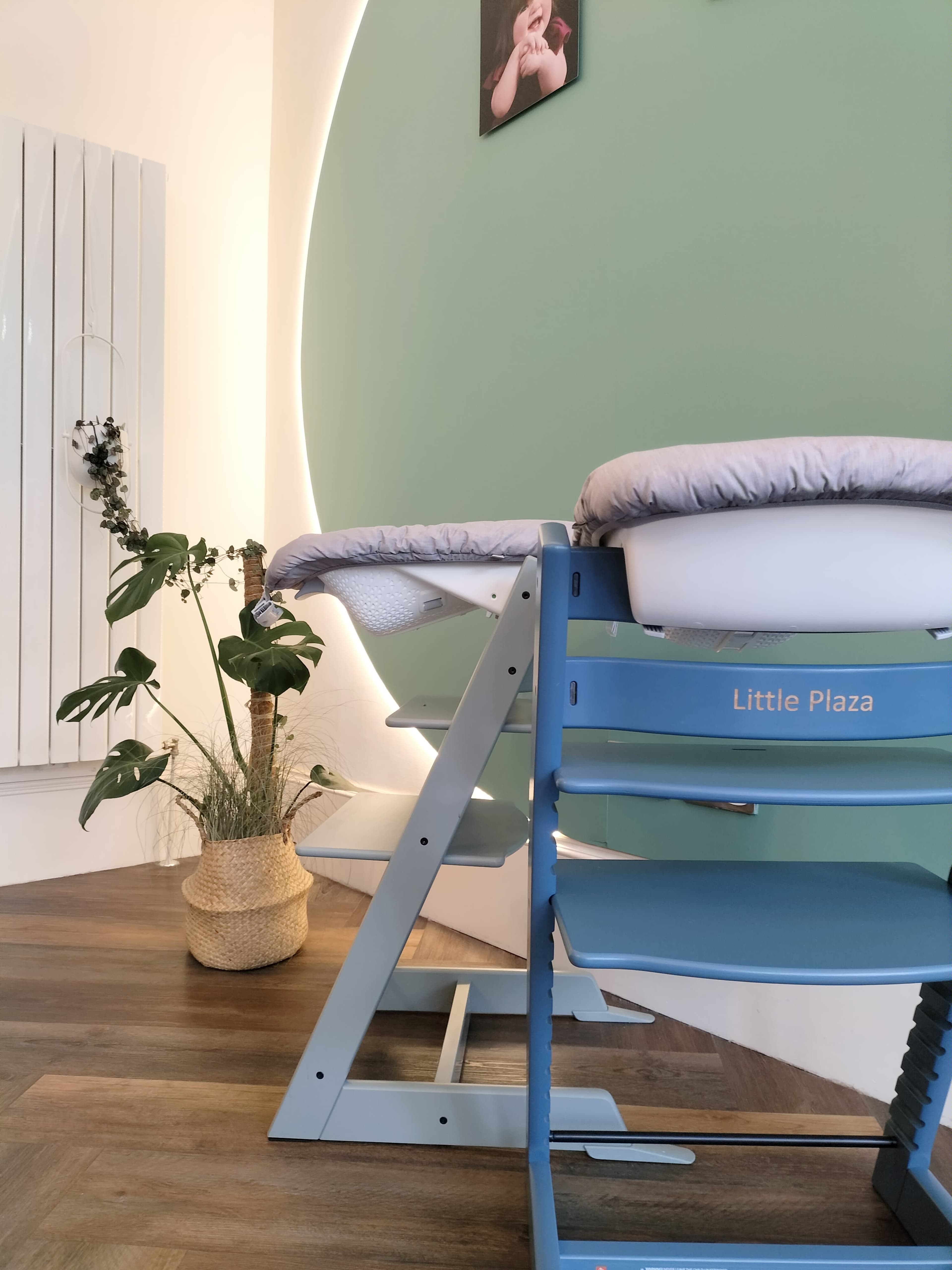 A wooden high chair labeled "Little Plaza" positioned next to a potted plant against a green wall with a circular light feature.