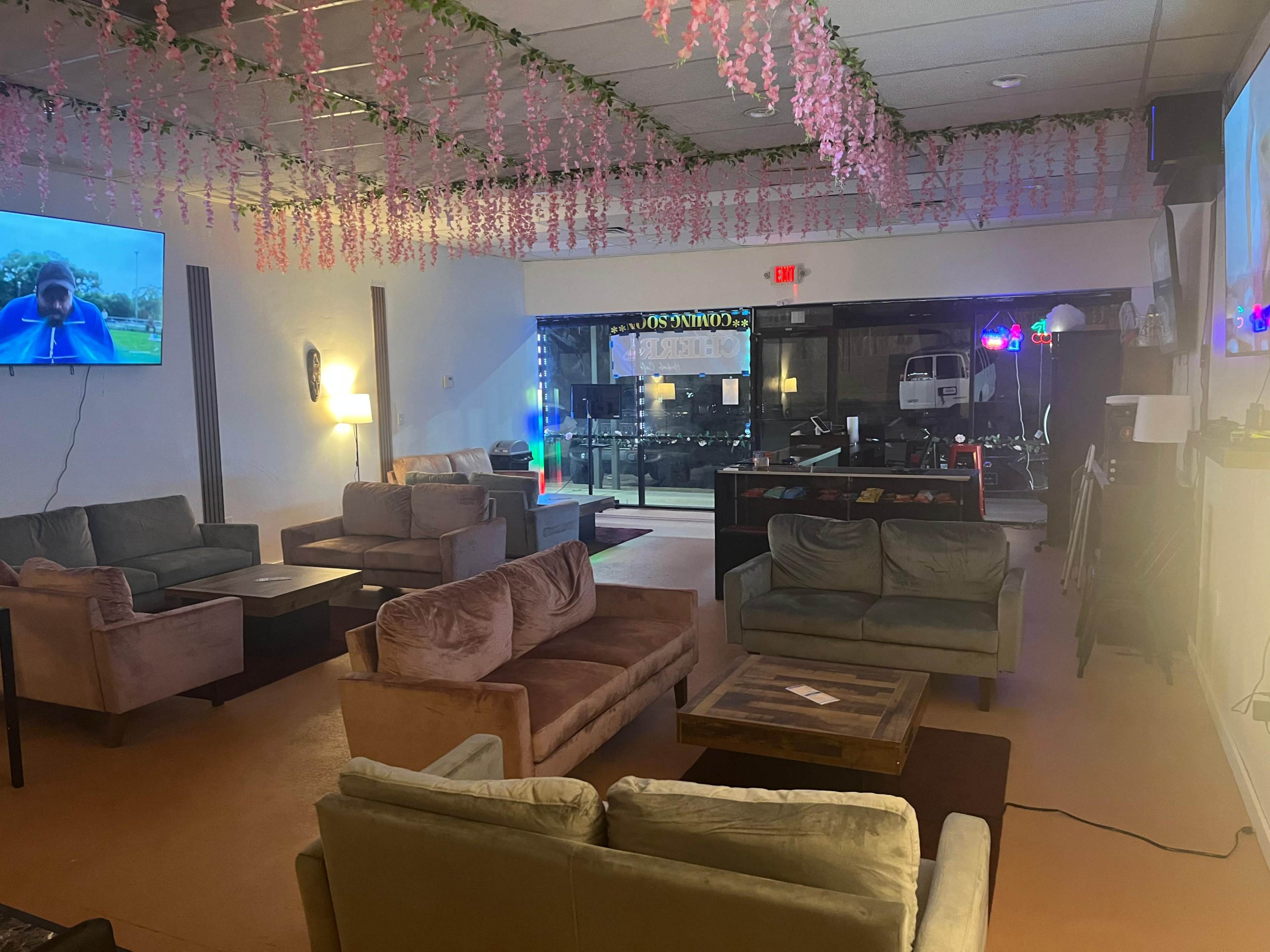 The image shows a cozy lounge area with several sofas arranged around a wooden coffee table, soft lighting, and floral decorations hanging from the ceiling.