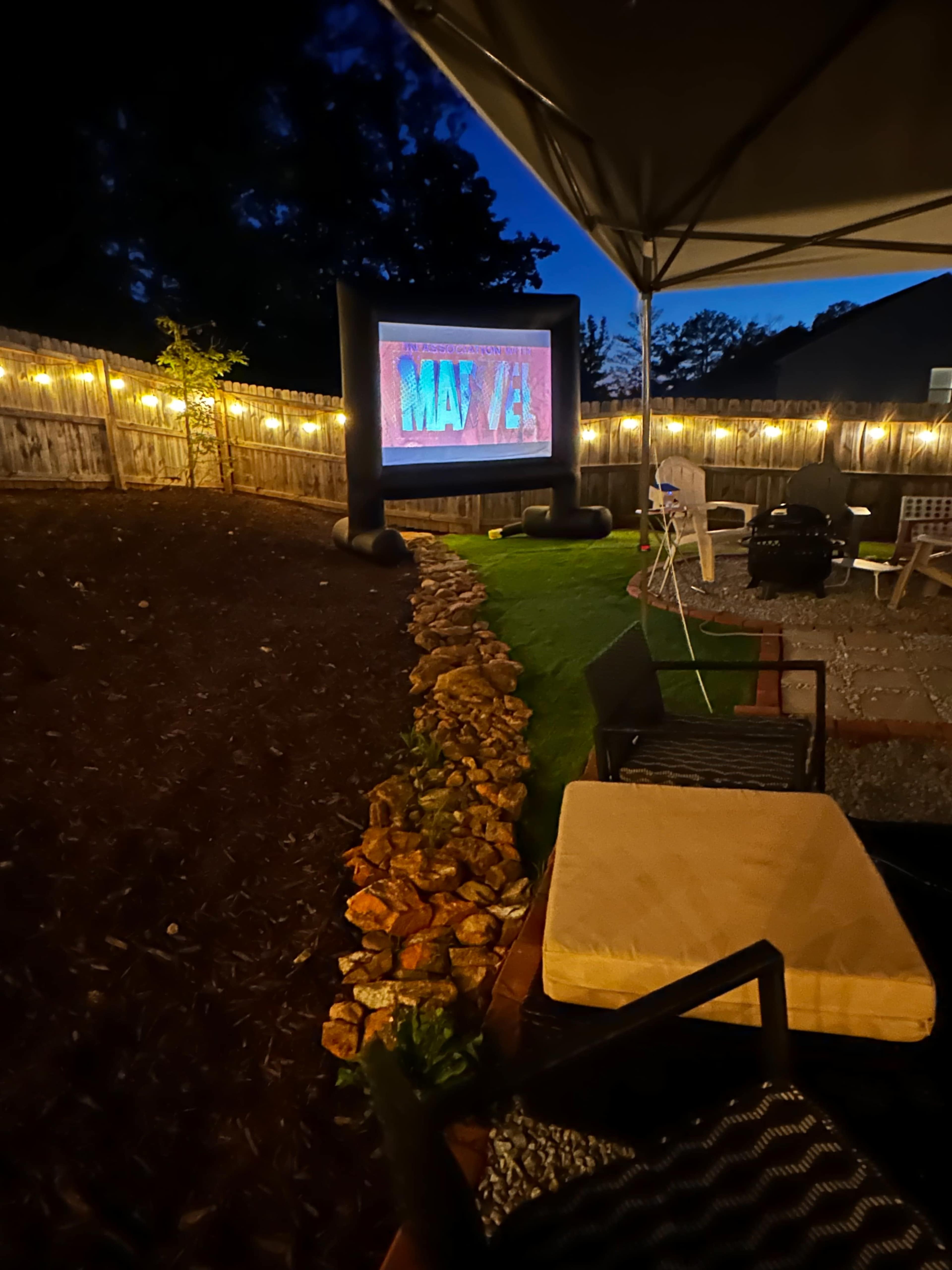 The scene shows an outdoor area at night with an inflatable movie screen displaying a colorful image, surrounded by string lights and seating arrangements.
