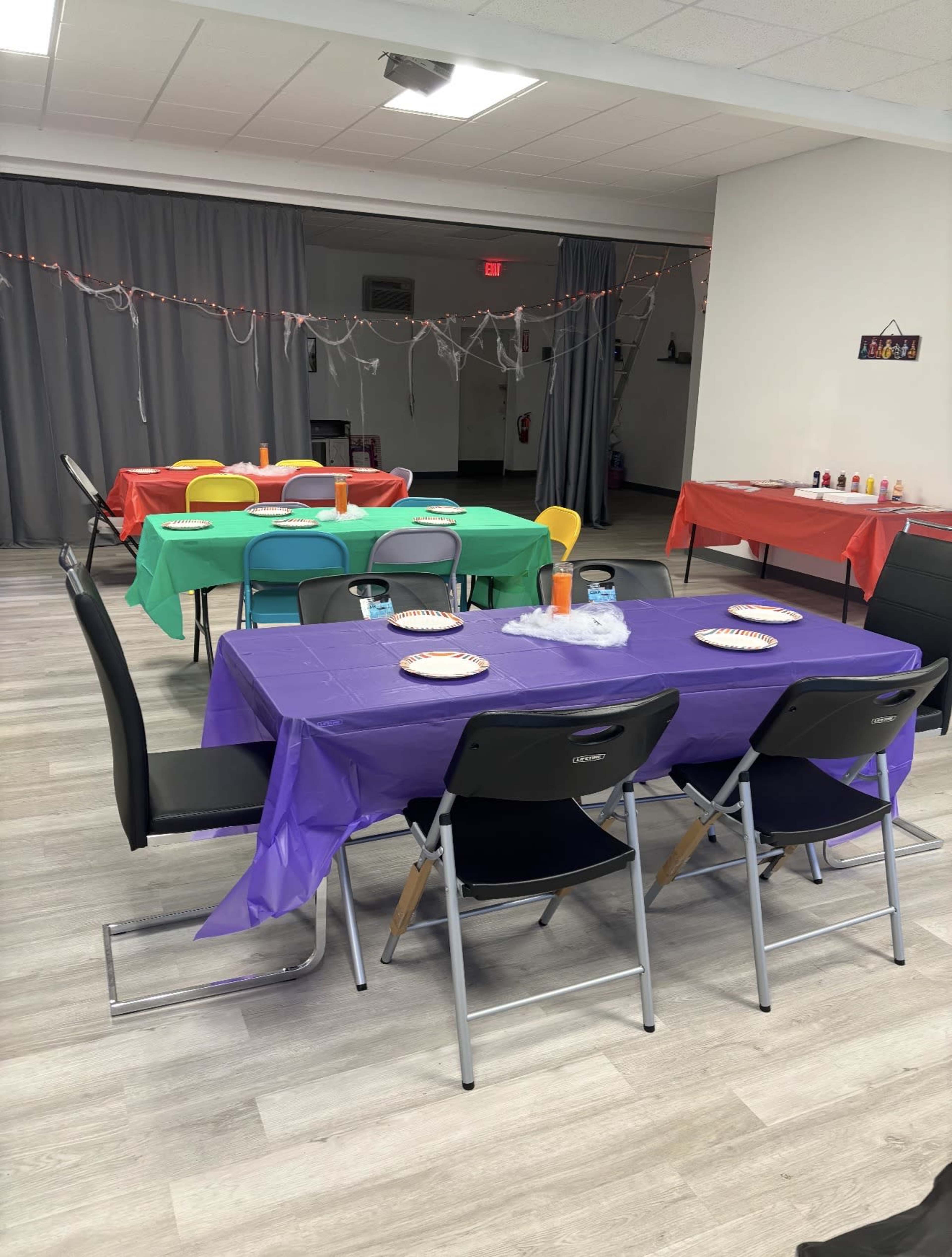 The image shows a decorated room with tables covered in colorful tablecloths and set up for a gathering, featuring plates and drinks on each table.