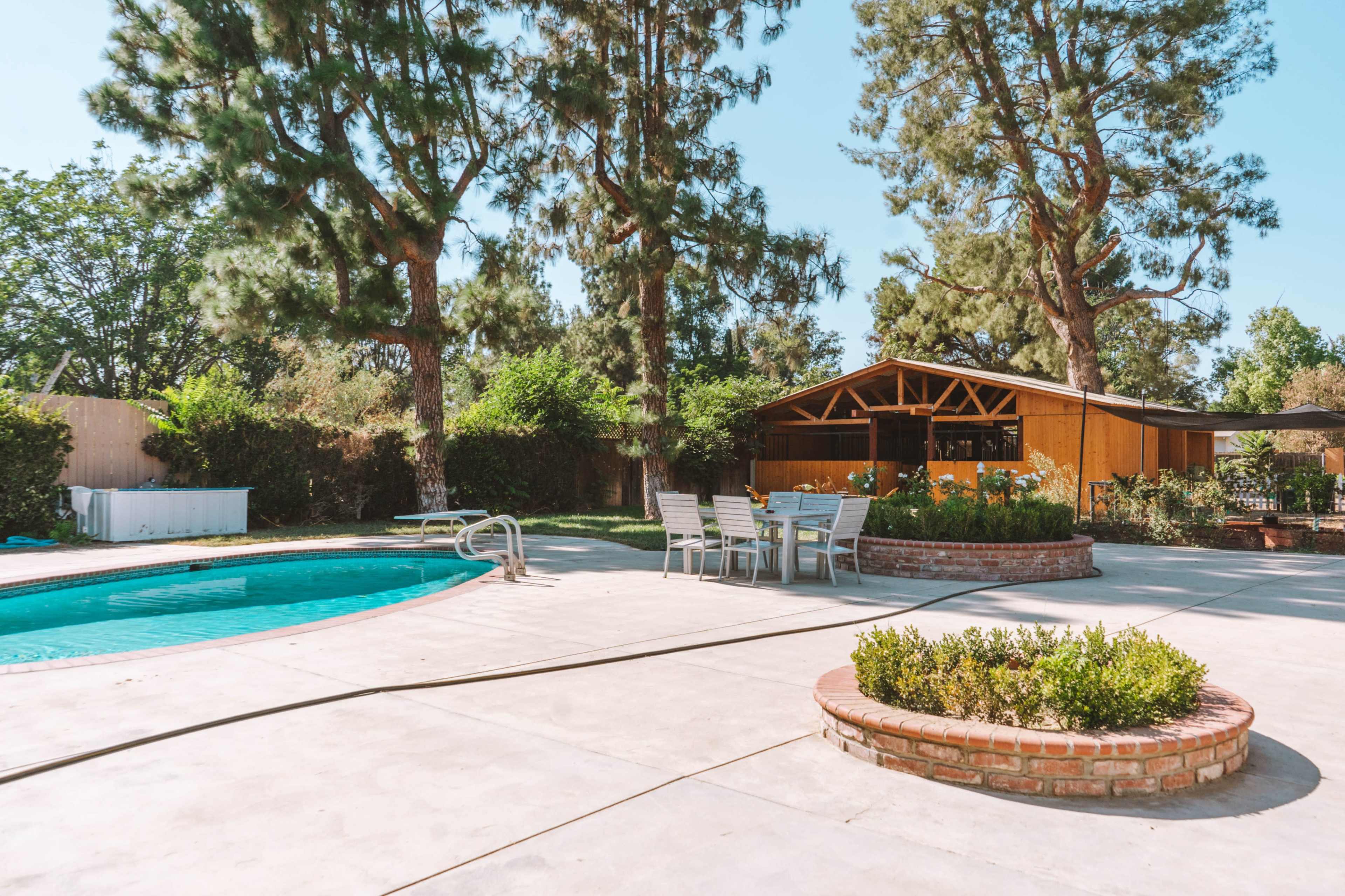 A swimming pool with a surrounding patio and seating area is situated next to a modern wooden house and tall pine trees.