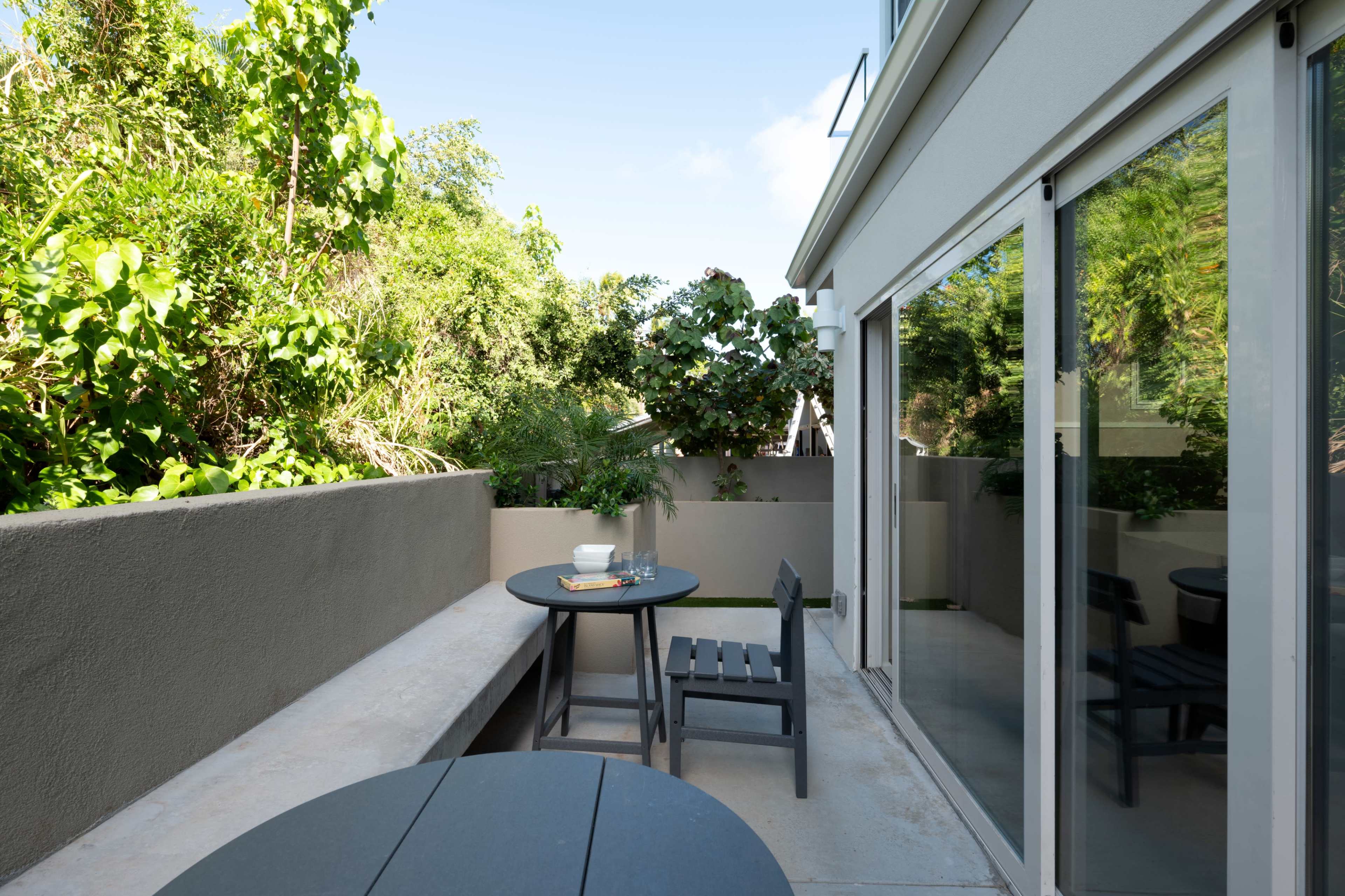 The image shows a small outdoor patio with two black tables and chairs surrounded by greenery.