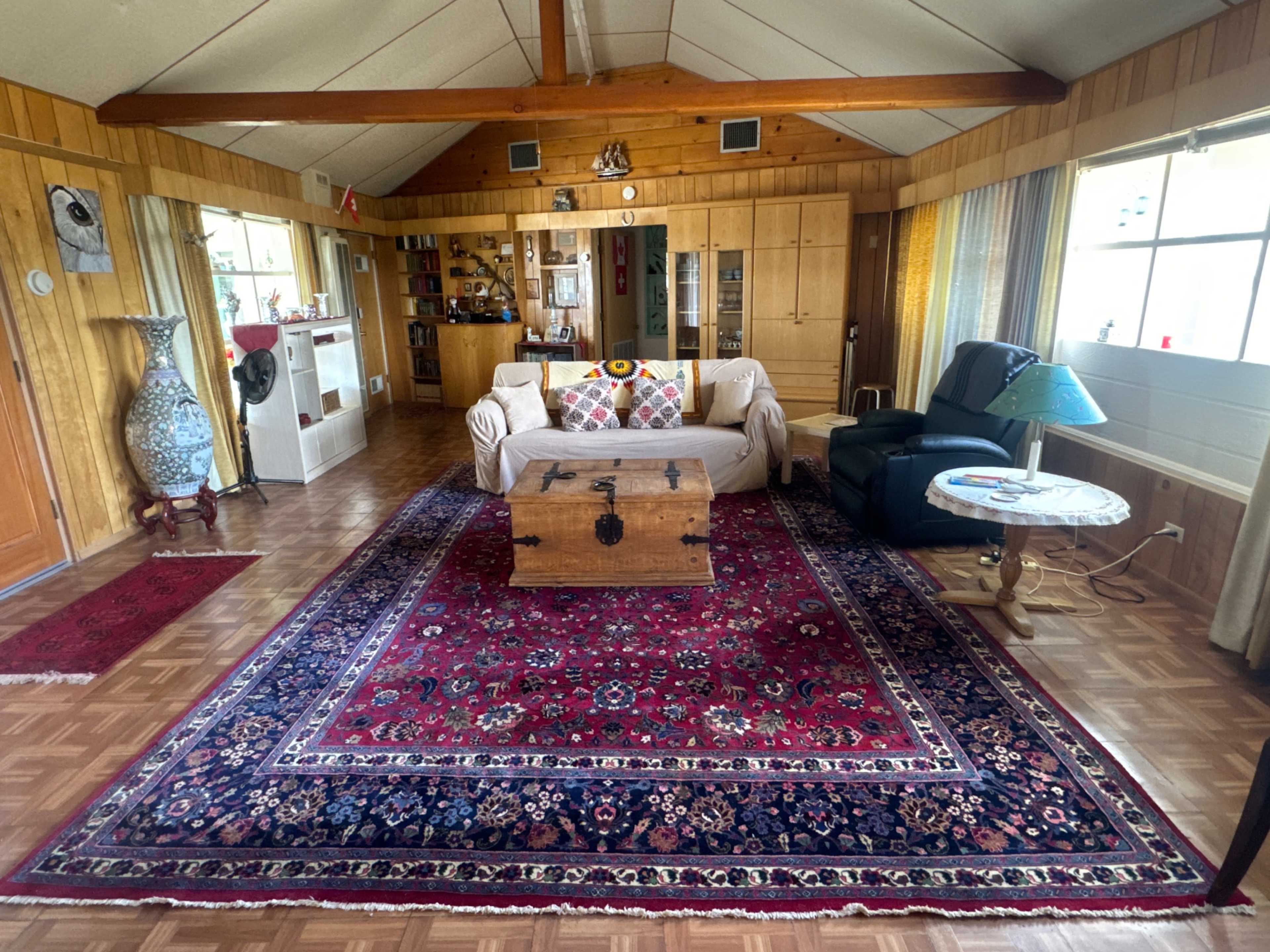 The living room features a patterned red rug, a wooden coffee table, a sofa with colorful pillows, and a green armchair, all set against wooden paneling and large windows.