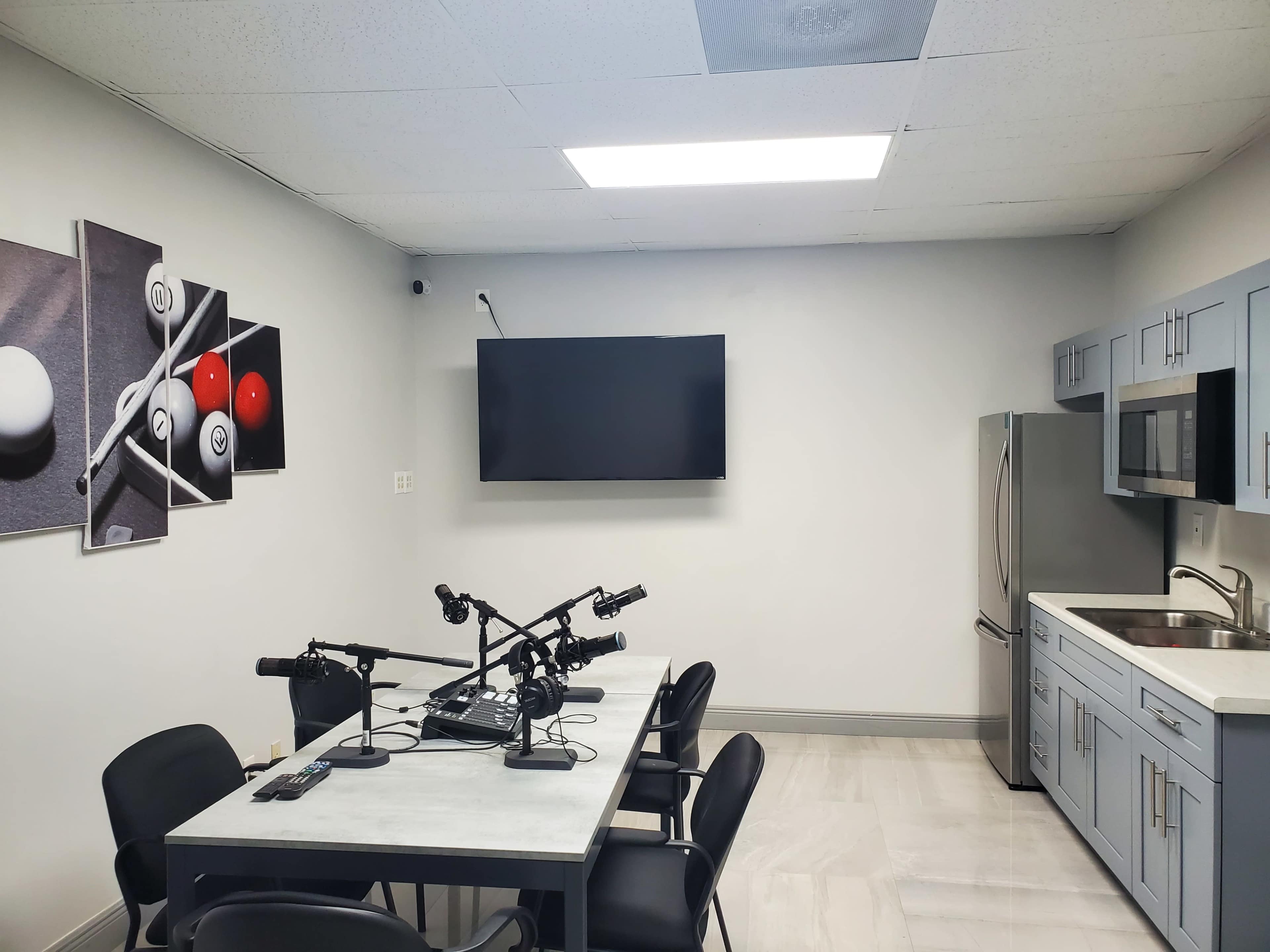 A modern kitchen space with a dining table equipped with microphones, a large wall-mounted television, and appliances including a refrigerator and microwave.