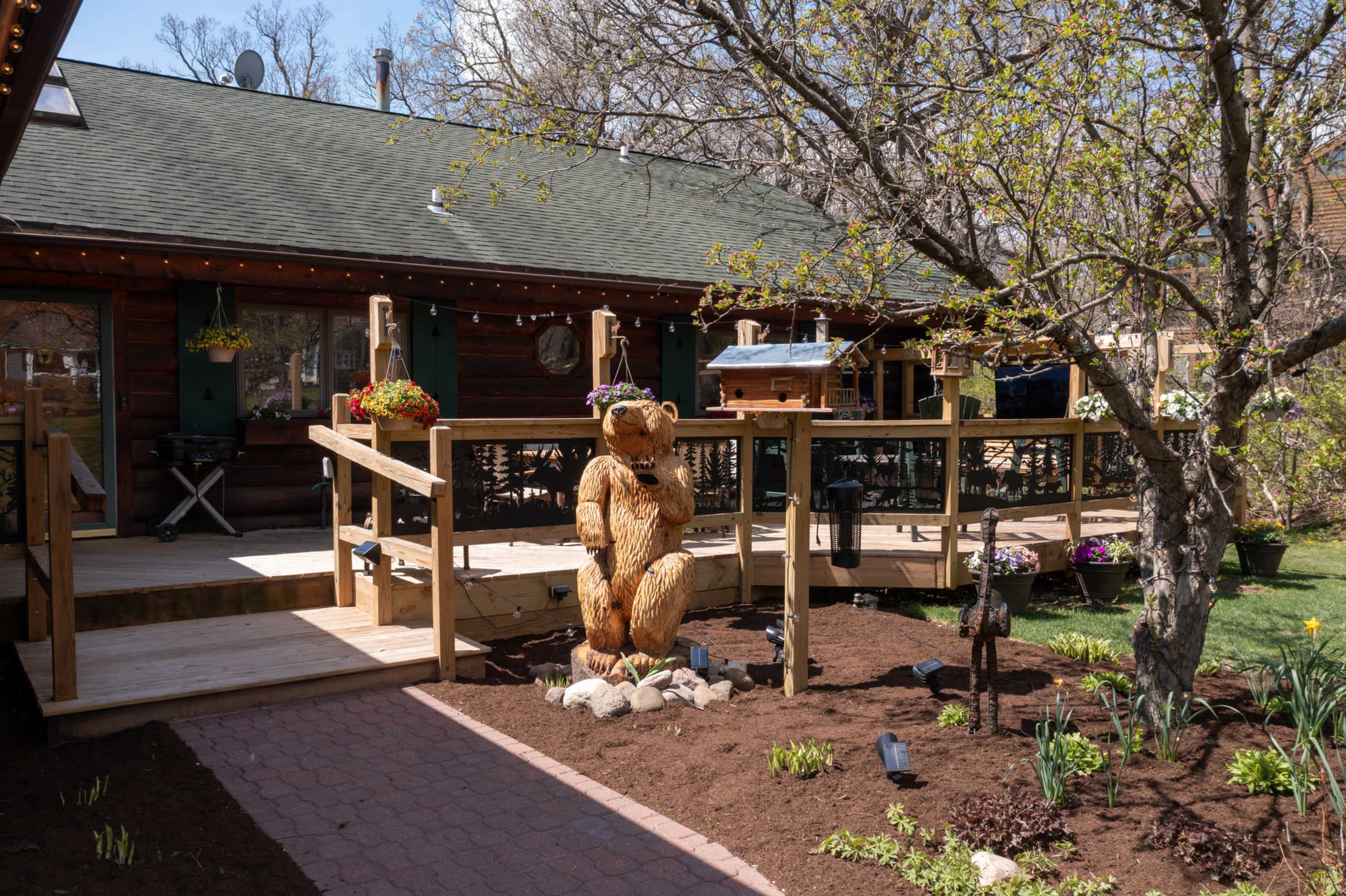 A wooden bear sculpture stands on the deck of a log cabin surrounded by a landscaped yard with flowering plants and a bird feeder.