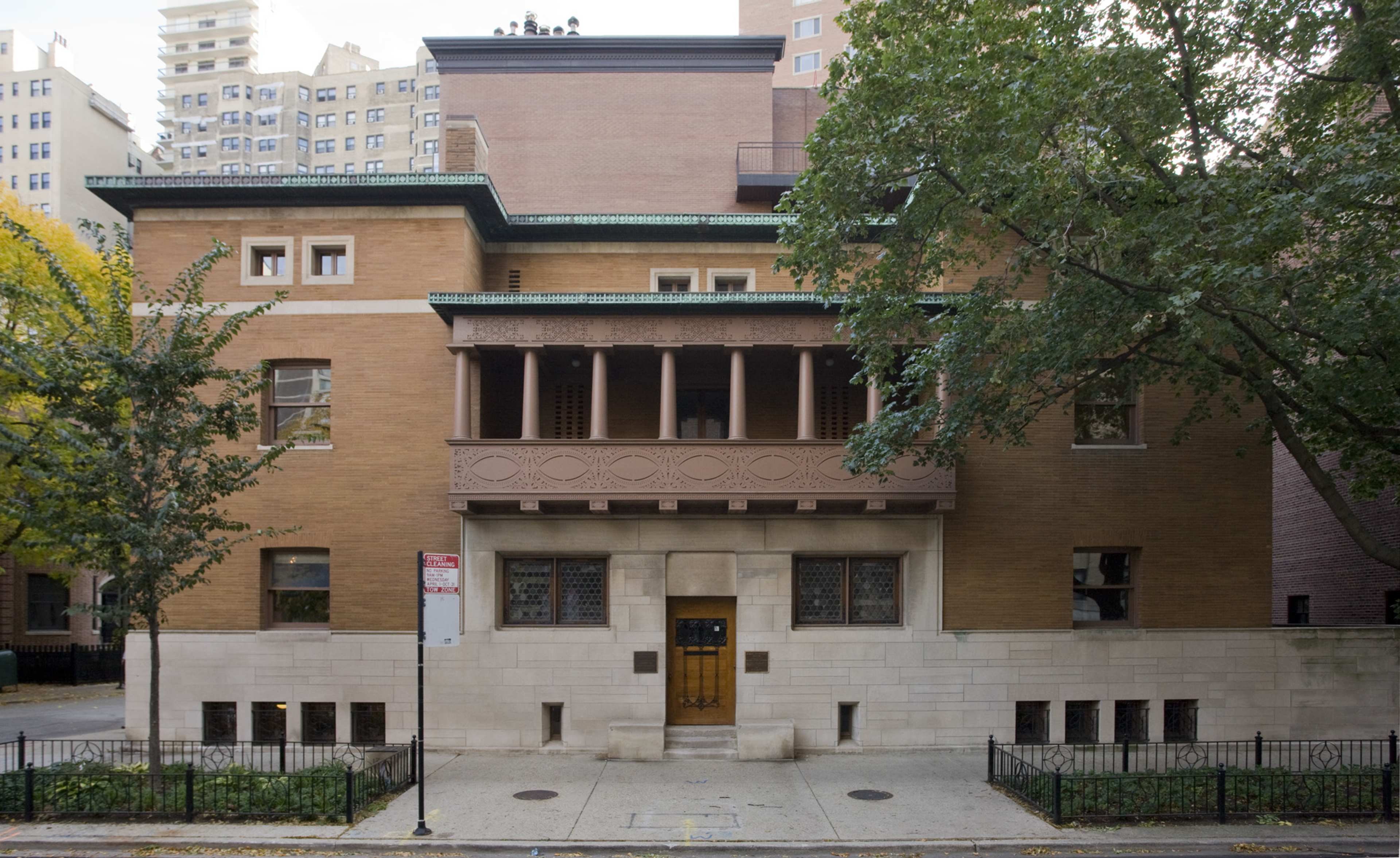 A large, two-story brick building with a distinctive overhanging balcony supported by columns, surrounded by trees and urban structures.