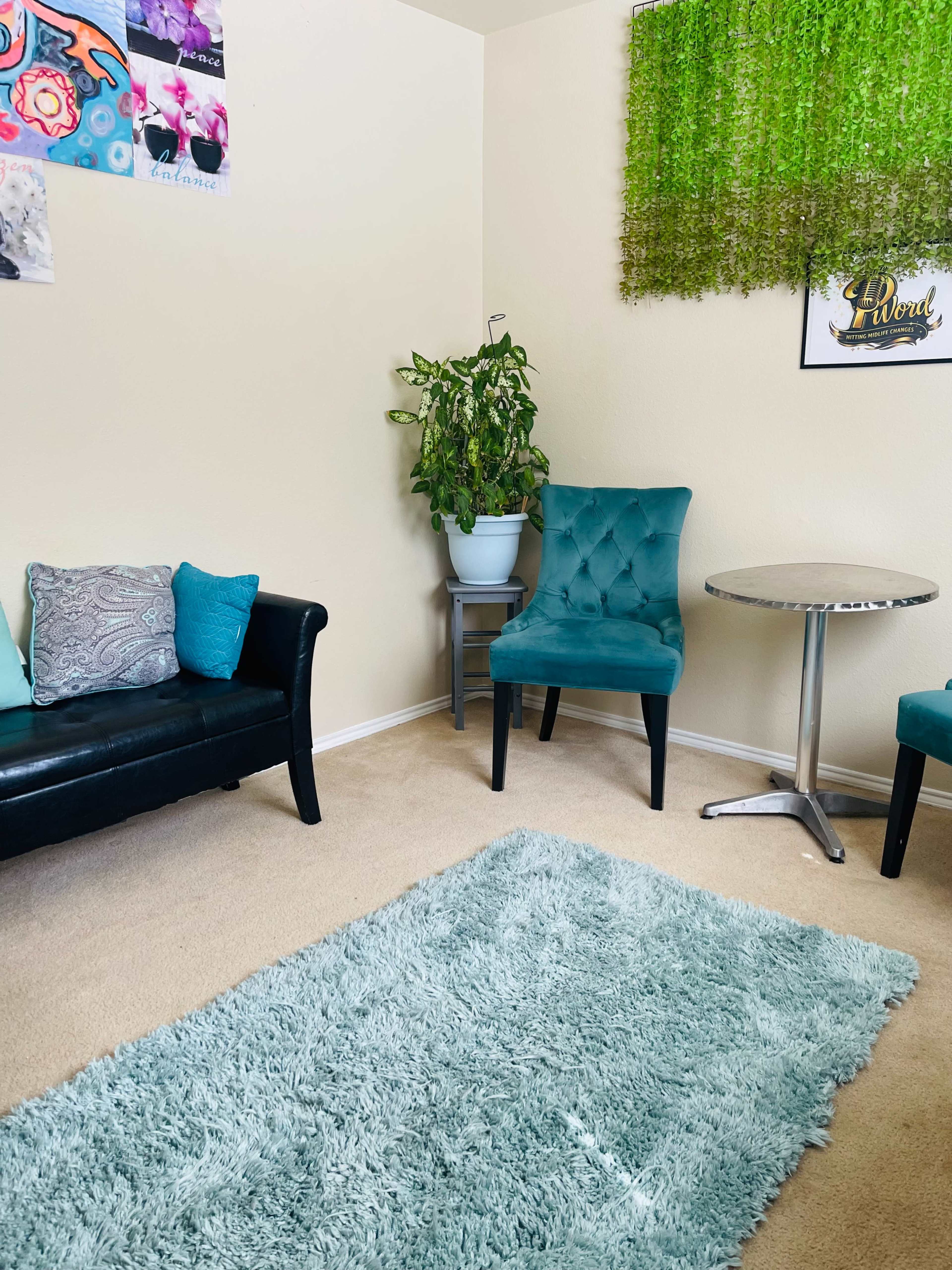 The image shows a cozy room featuring a black couch with turquoise pillows, a green plant in a pot, two blue chairs, a small round table, and a textured rug on the carpeted floor.