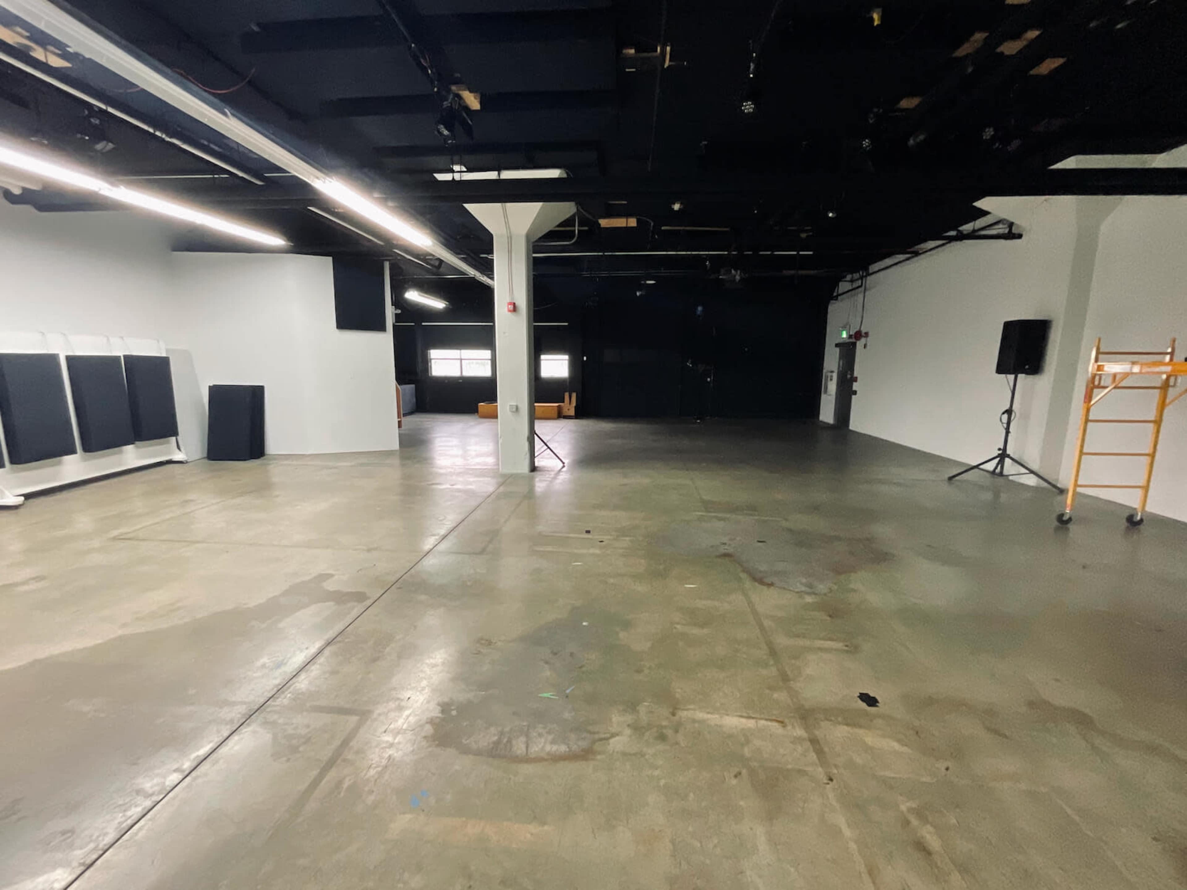 The image shows a large, empty industrial space with concrete flooring, exposed ceiling, and minimal furnishings.