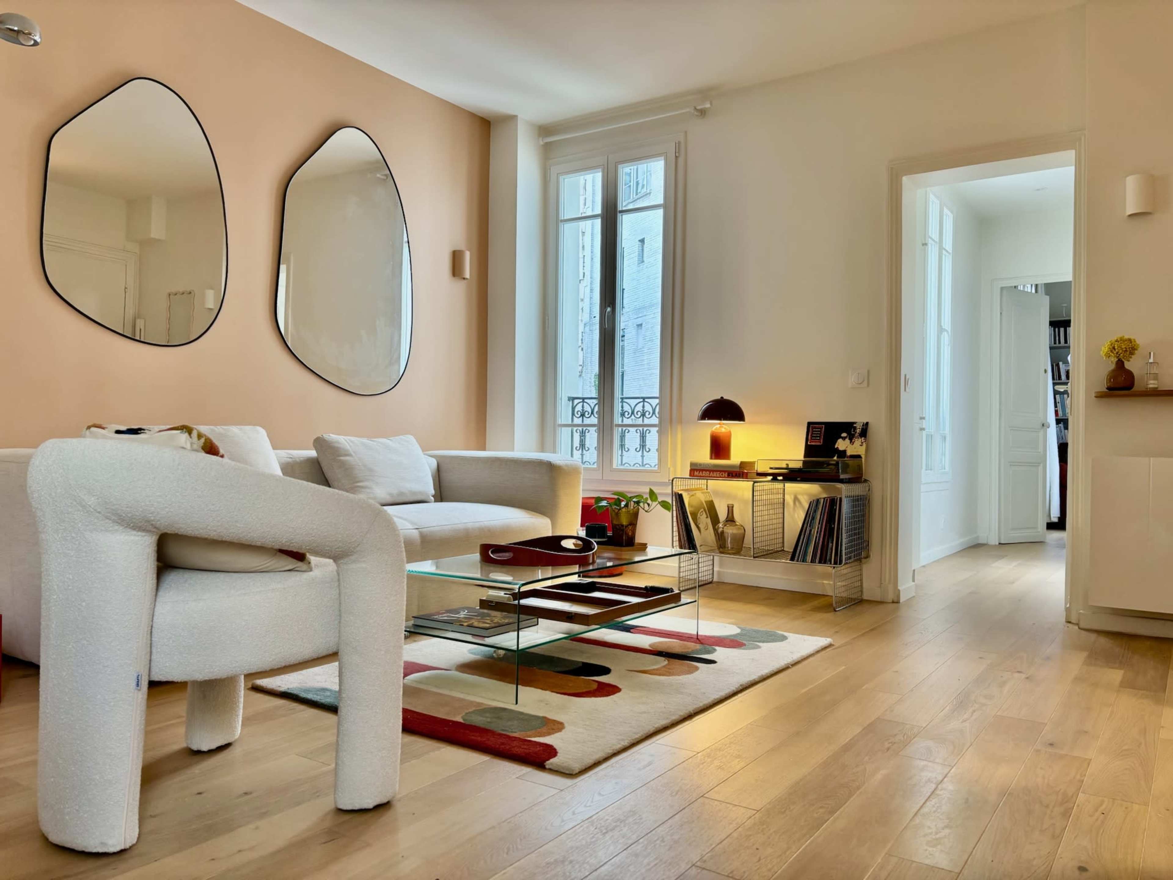 A modern living room features a light-colored sofa, unique-shaped mirrors, a glass coffee table, and a blend of minimalistic decor and natural light.