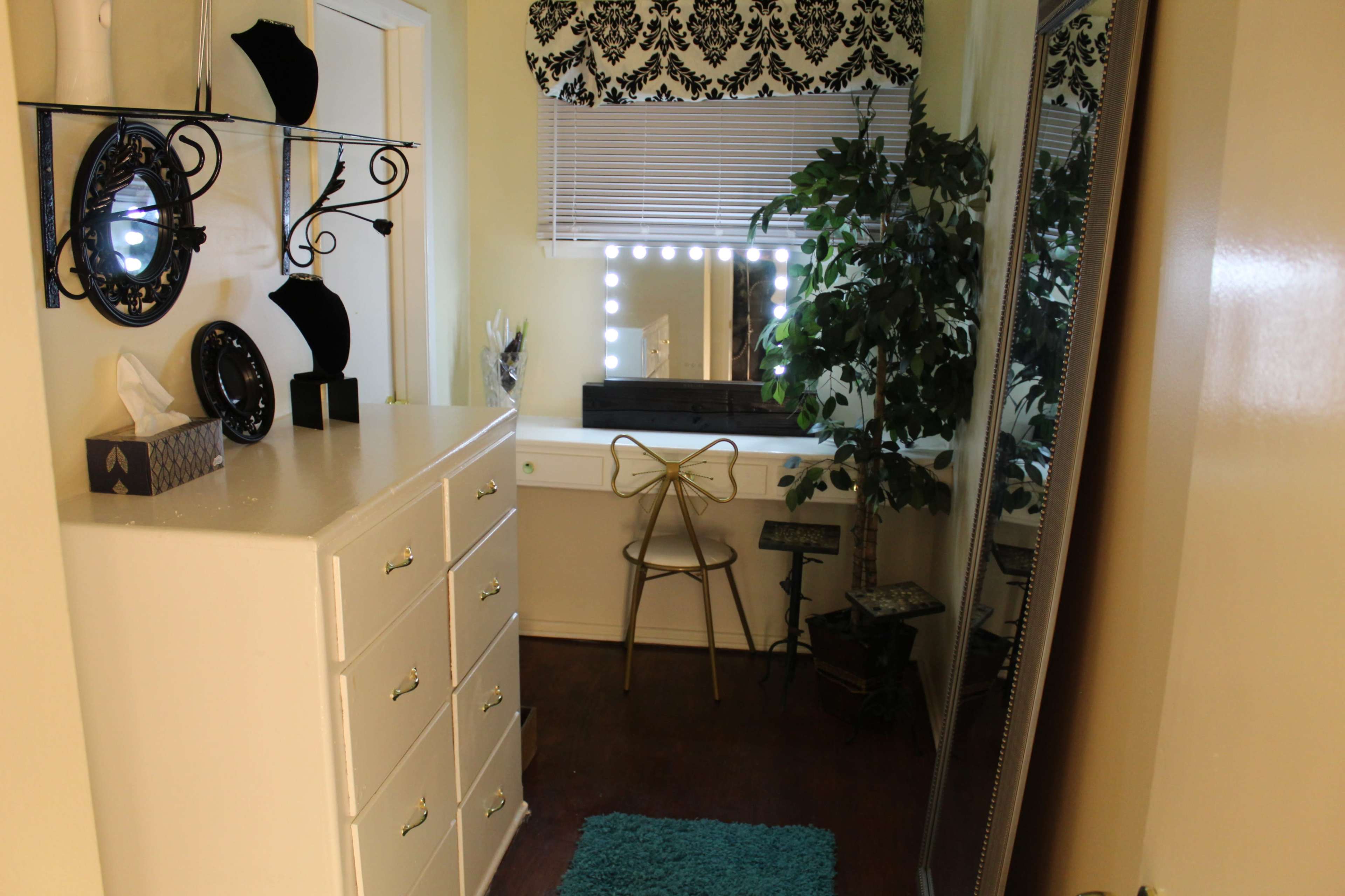 The image shows a small dressing area with a mirrored vanity table, a chair, a decorative plant, and a dresser with multiple drawers.