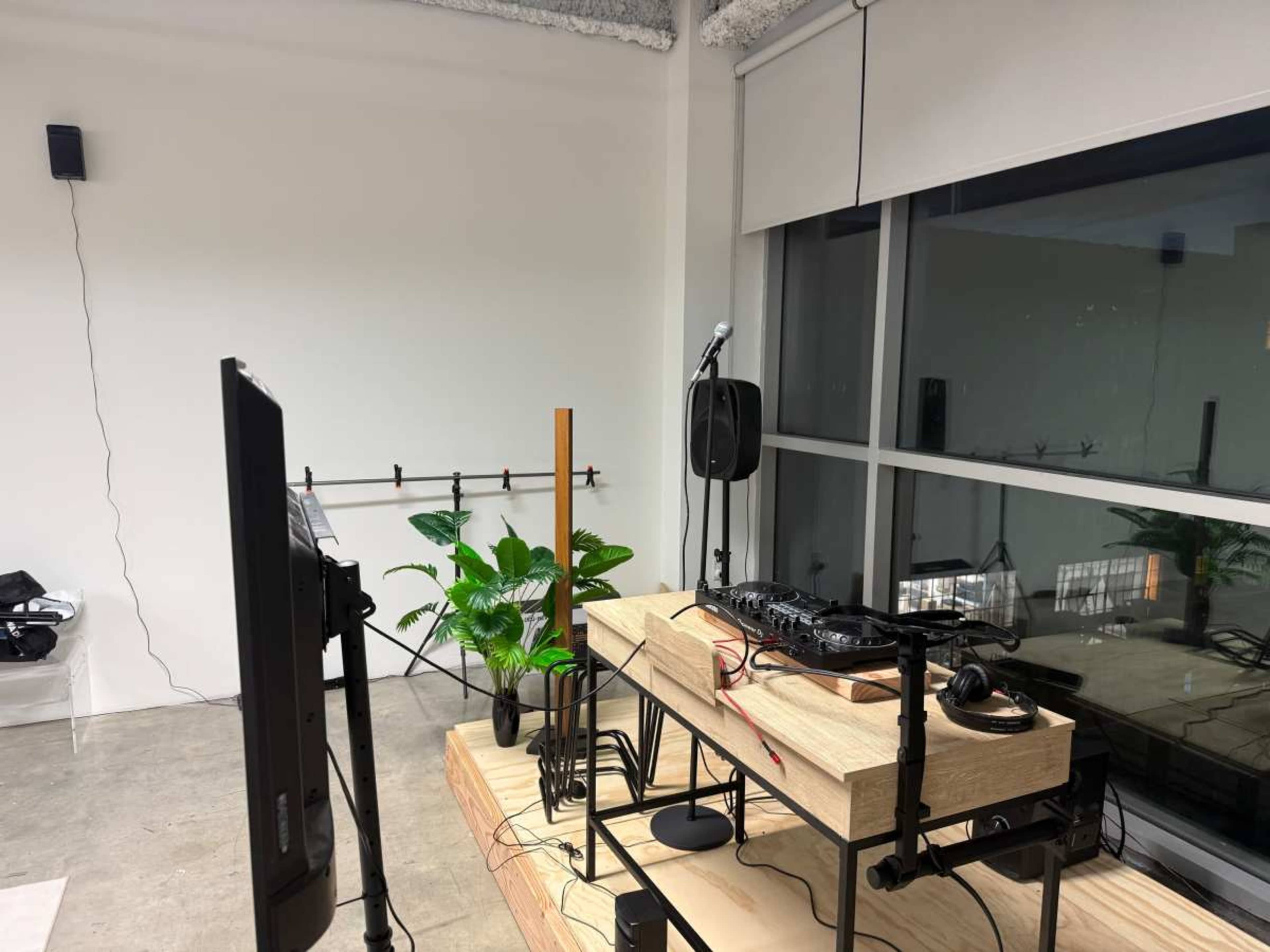 The image shows a DJ setup with a mixer and turntables placed on a wooden table, accompanied by a microphone and speakers in a bright room with large windows and plants.