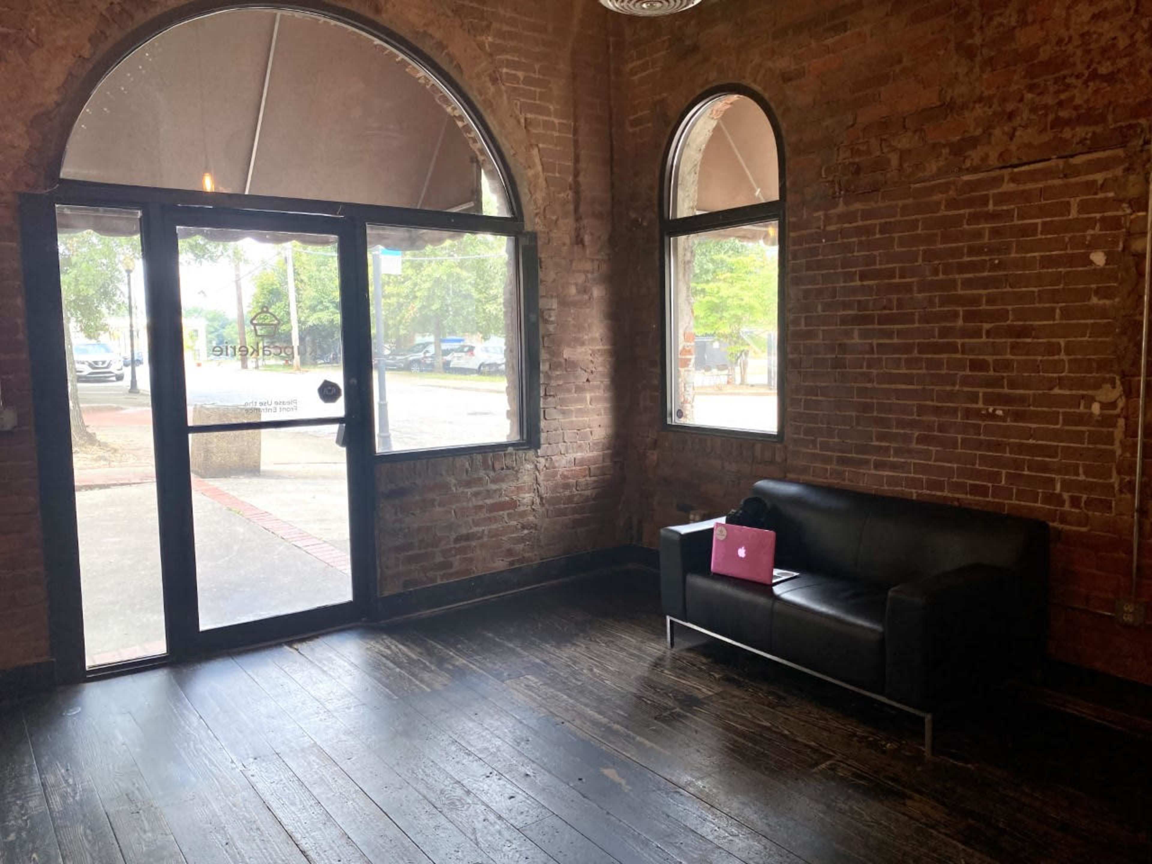 A waiting area with a black couch and a closed glass door, framed by brick walls and large windows.