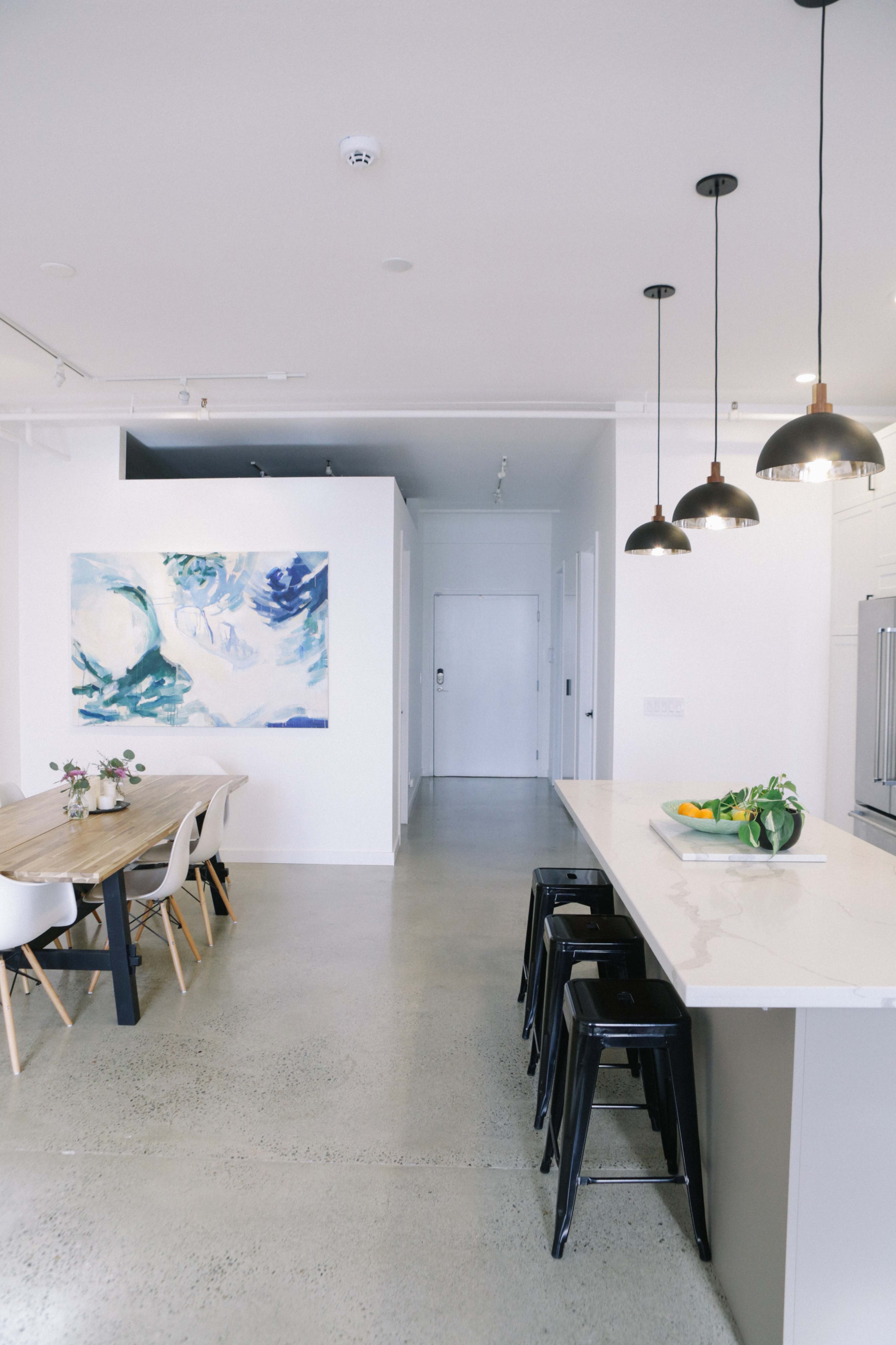 A modern kitchen and dining area features a long table, high stools, and a large abstract painting on the wall.