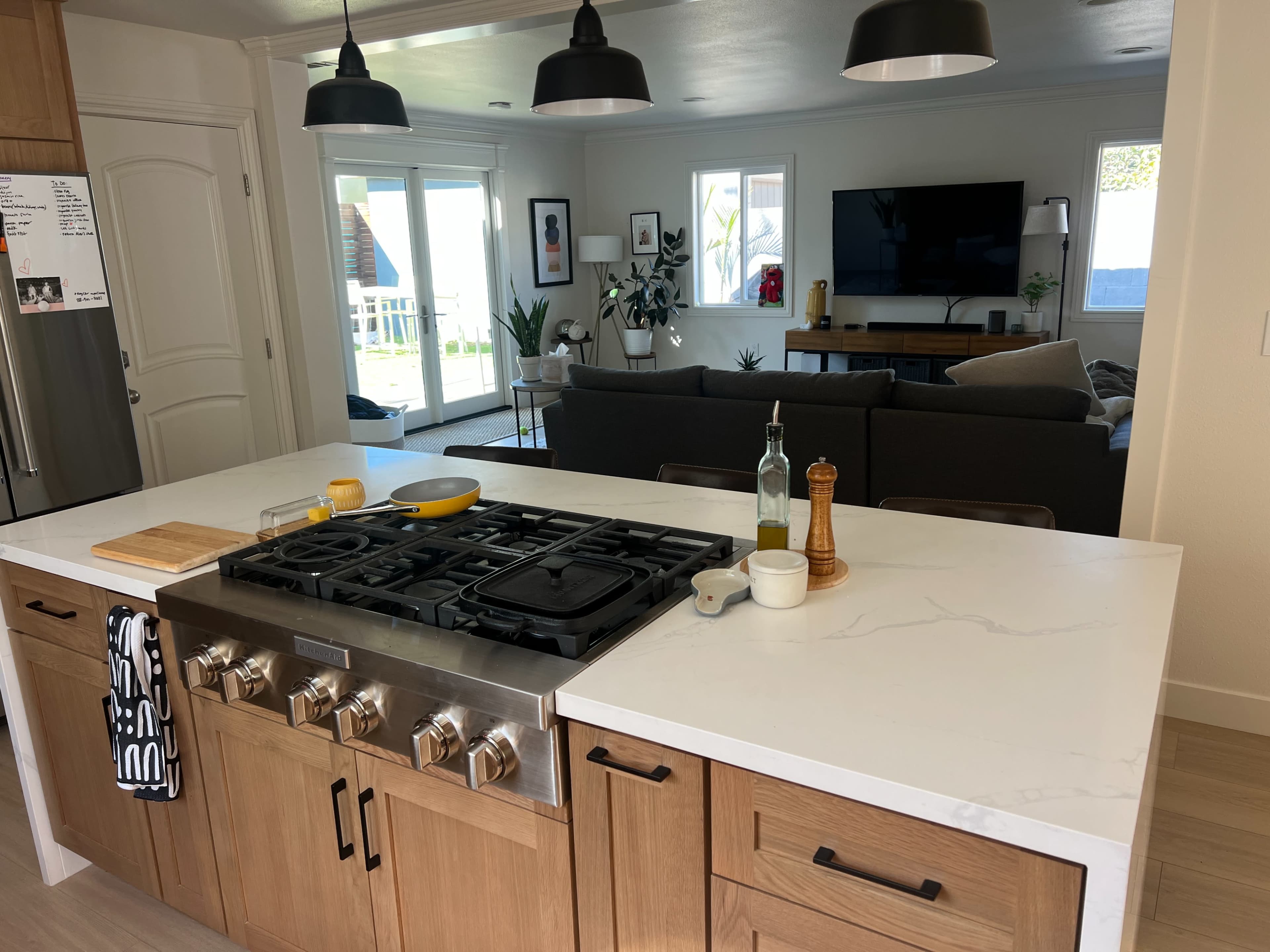 A modern kitchen features wooden cabinetry, a gas stove with a sleek countertop, and an open layout leading to a living area with a television and seating.