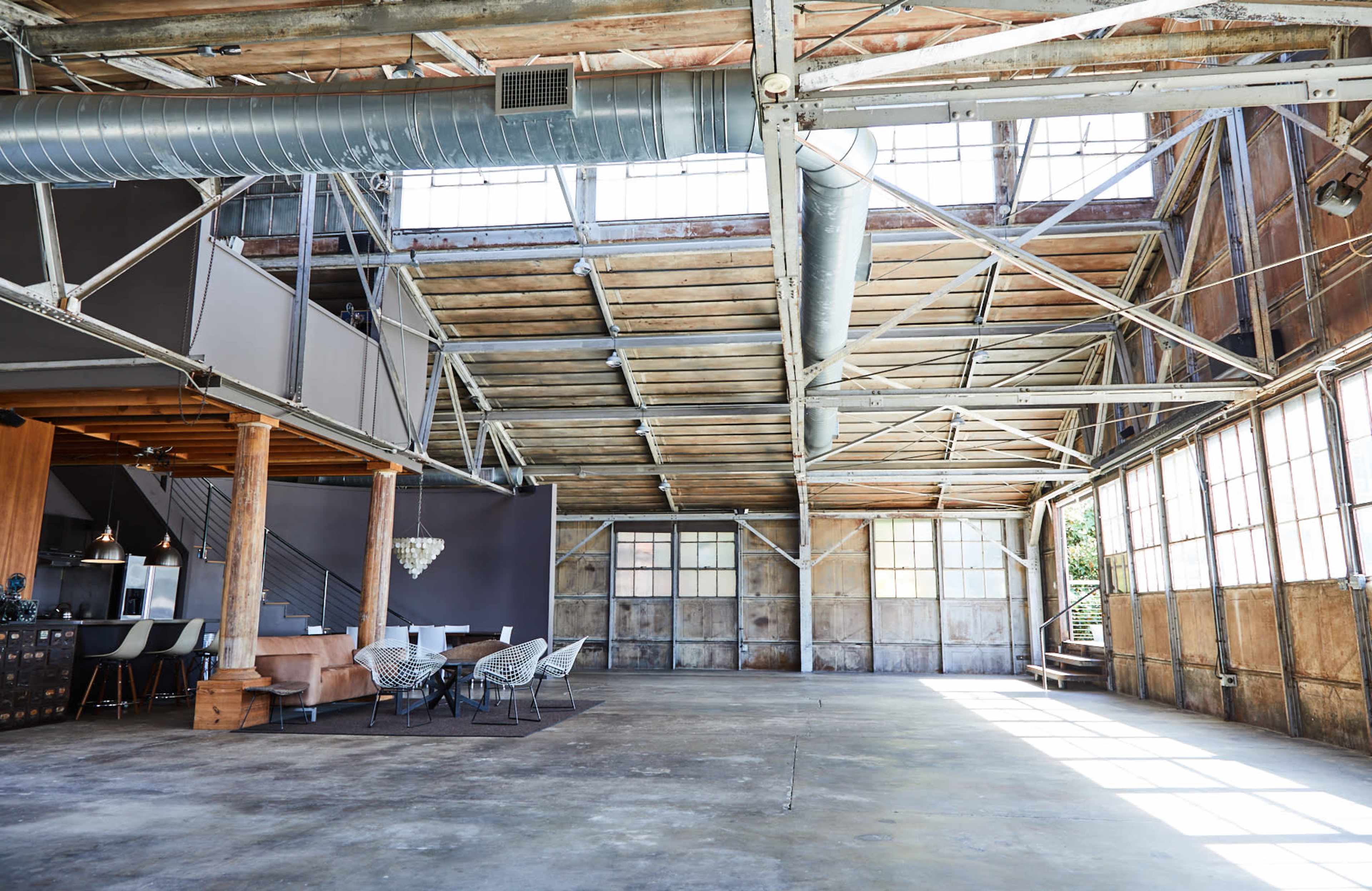The image shows a spacious industrial-style interior with high ceilings, large windows, and minimal furnishings.