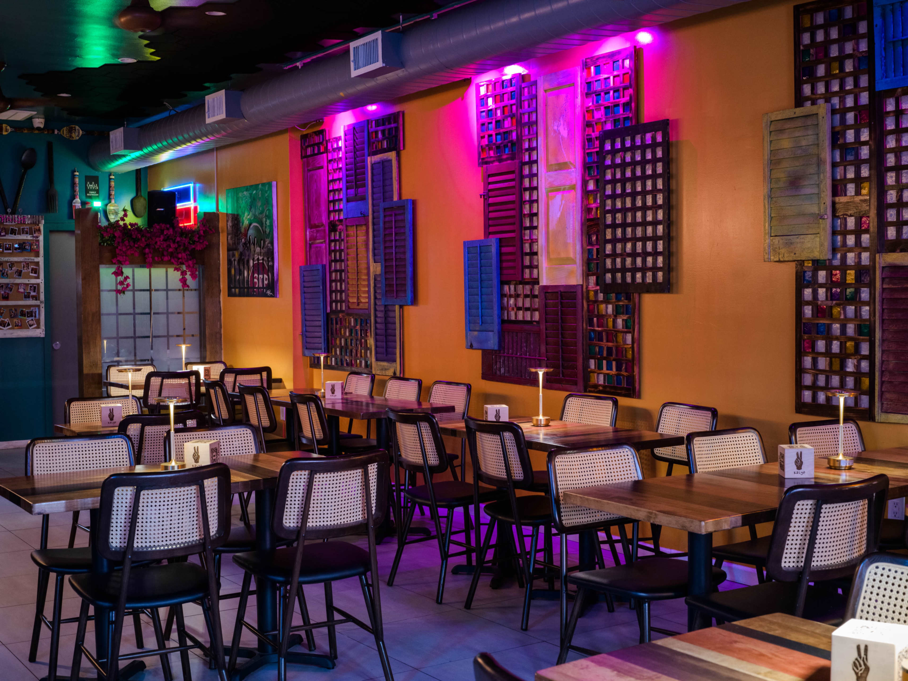 A colorful restaurant interior features wooden tables and black chairs against a wall adorned with vibrant, patterned decorations and ambient lighting.
