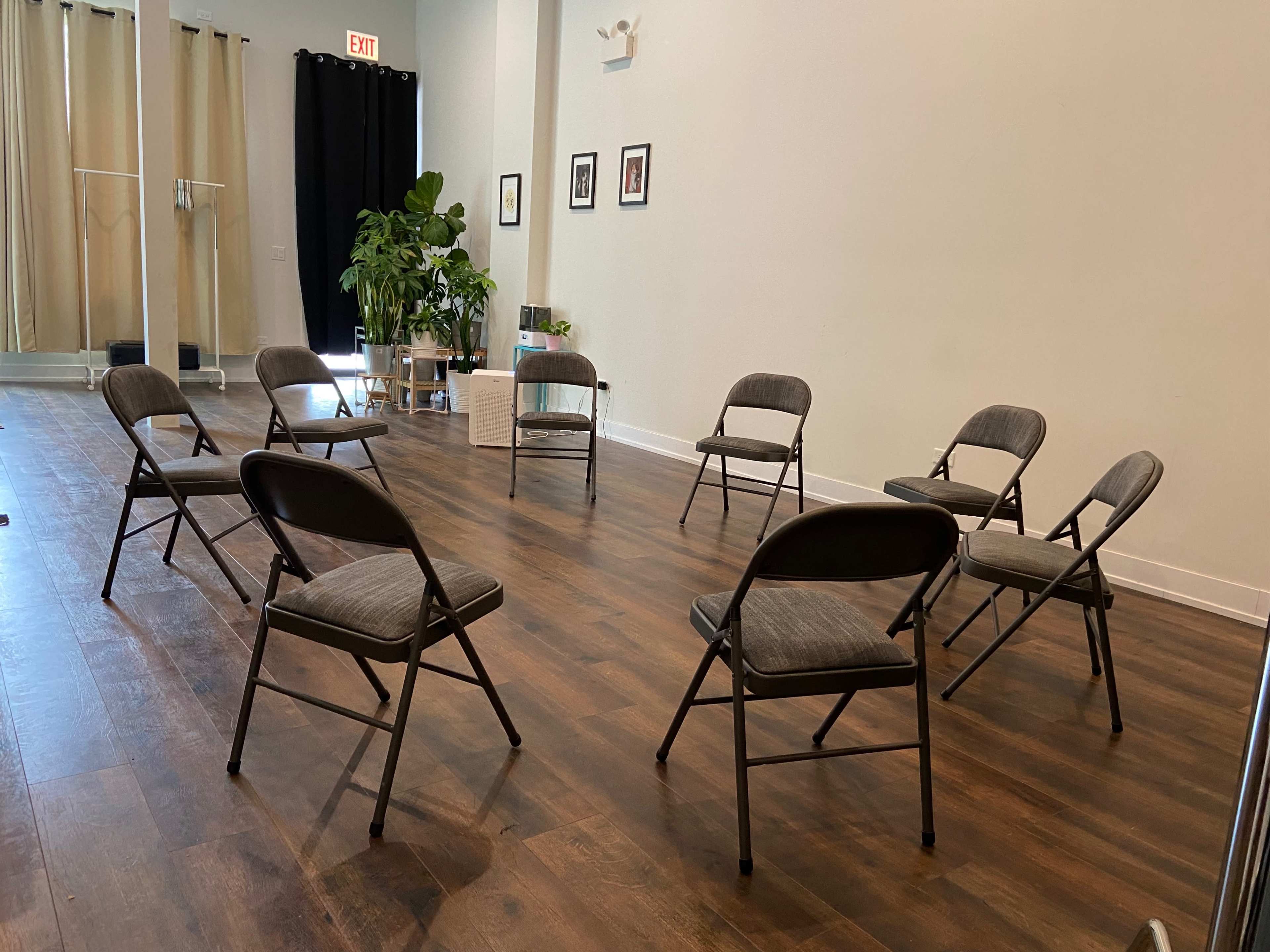 A circle of metal folding chairs is arranged on a wooden floor in a bright room with a large plant in the corner and light-colored curtains.