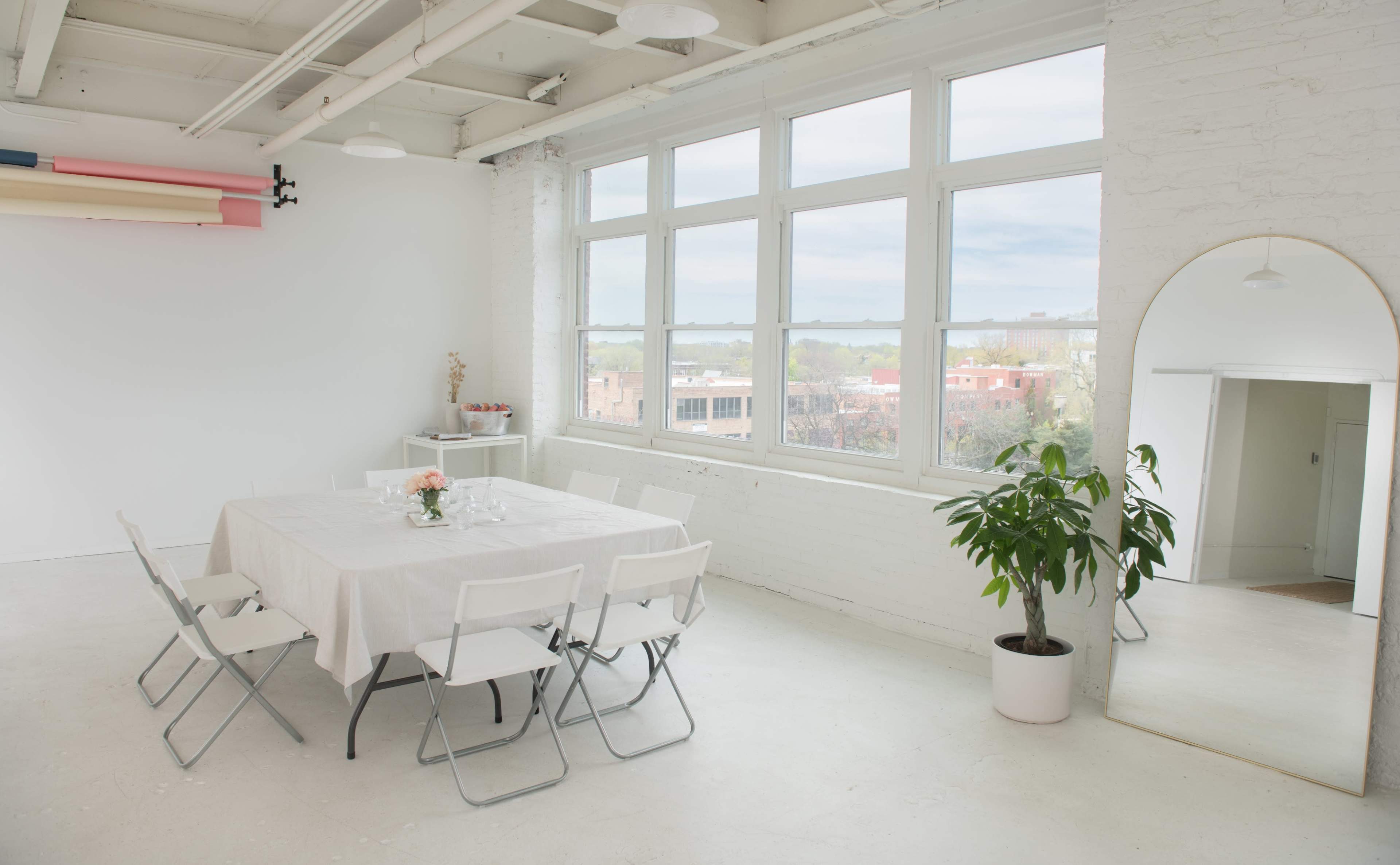 A bright, minimalistic room features a white table set for dining, surrounded by folding chairs, with large windows allowing natural light to enter.