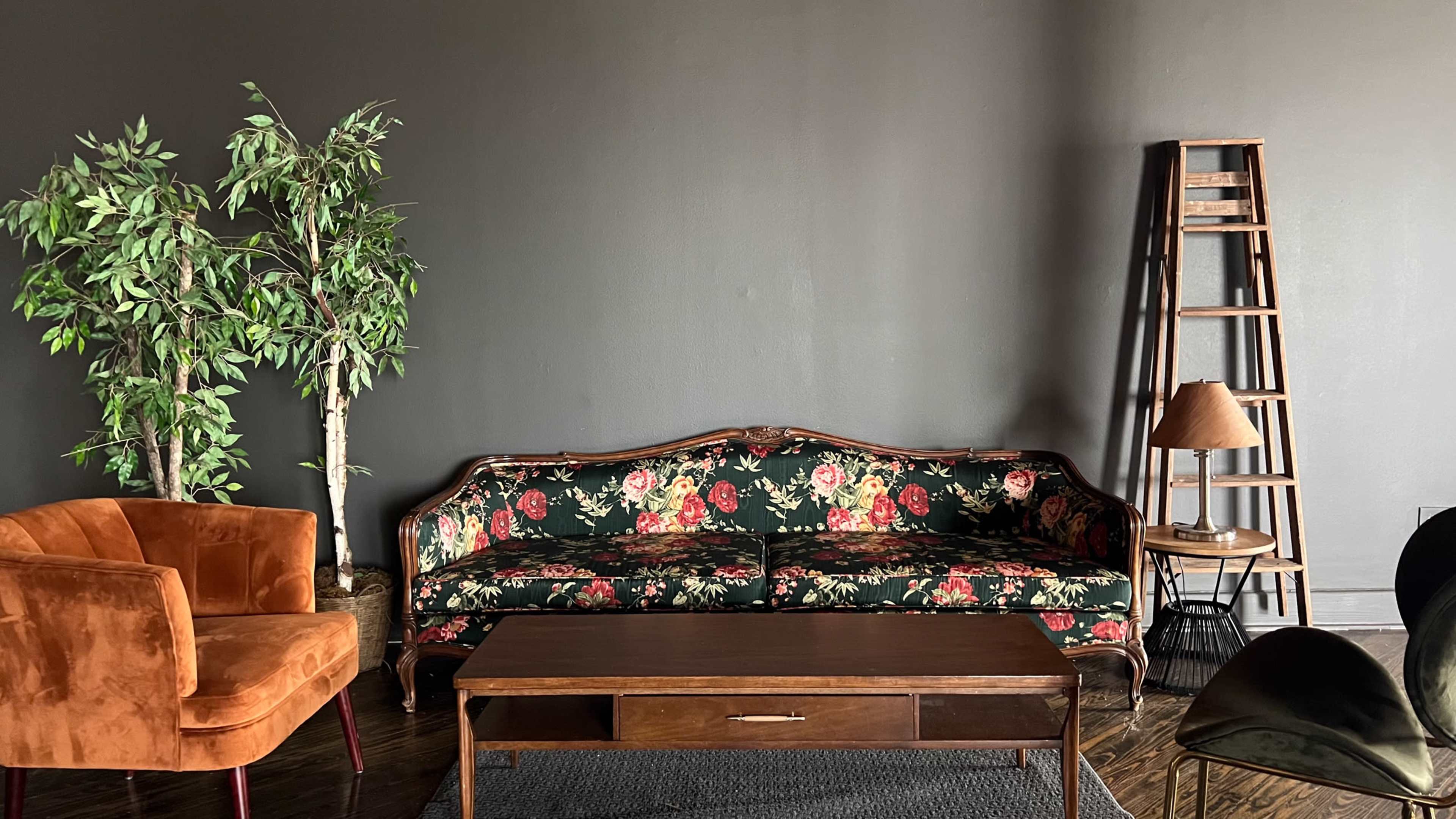A living room with a floral-patterned sofa, an orange armchair, a wooden coffee table, a floor lamp, and a tall wooden ladder against a dark wall.