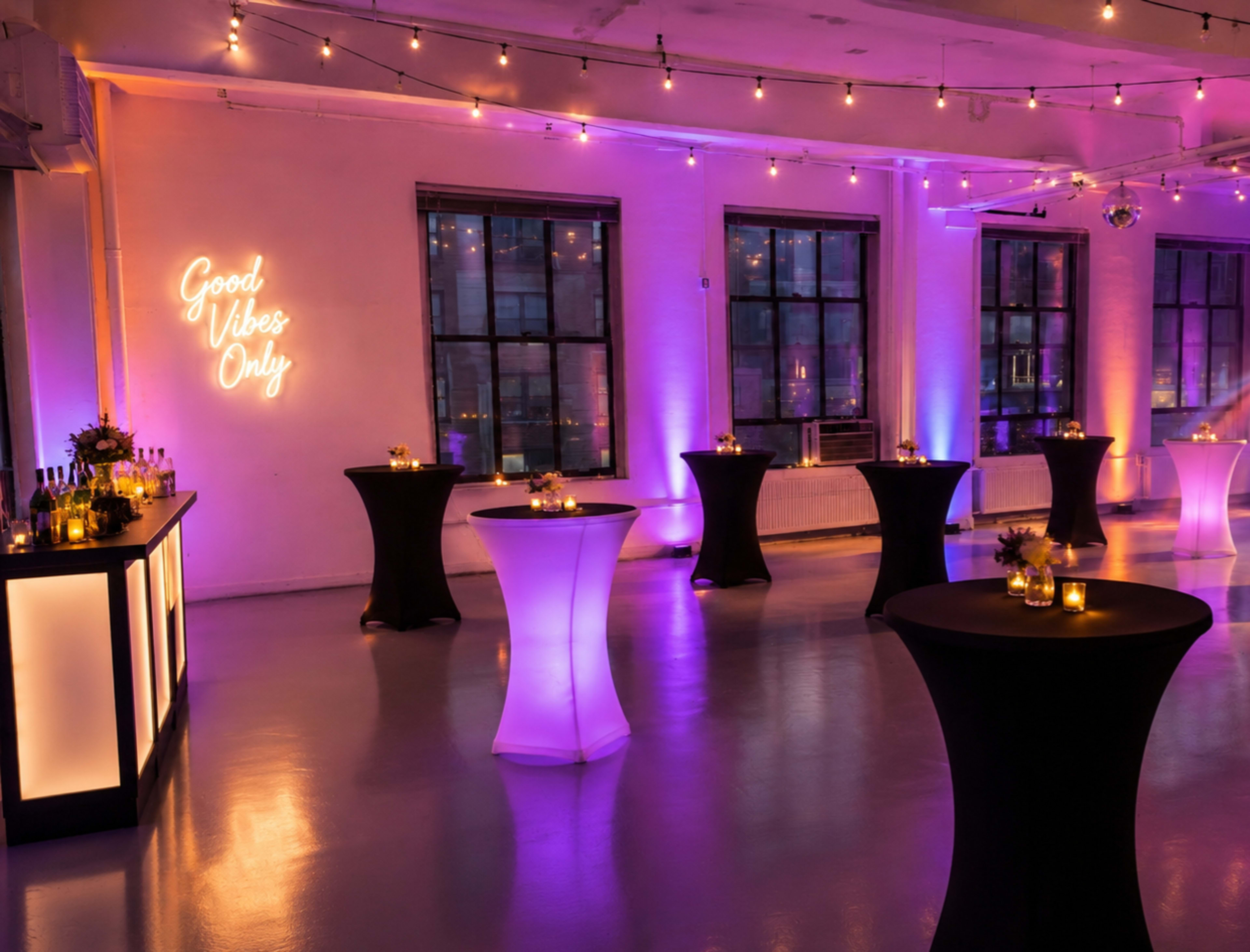 The image shows a decorated event space with tables, a bar, and ambient lighting featuring a neon sign that reads "Good Vibes Only."