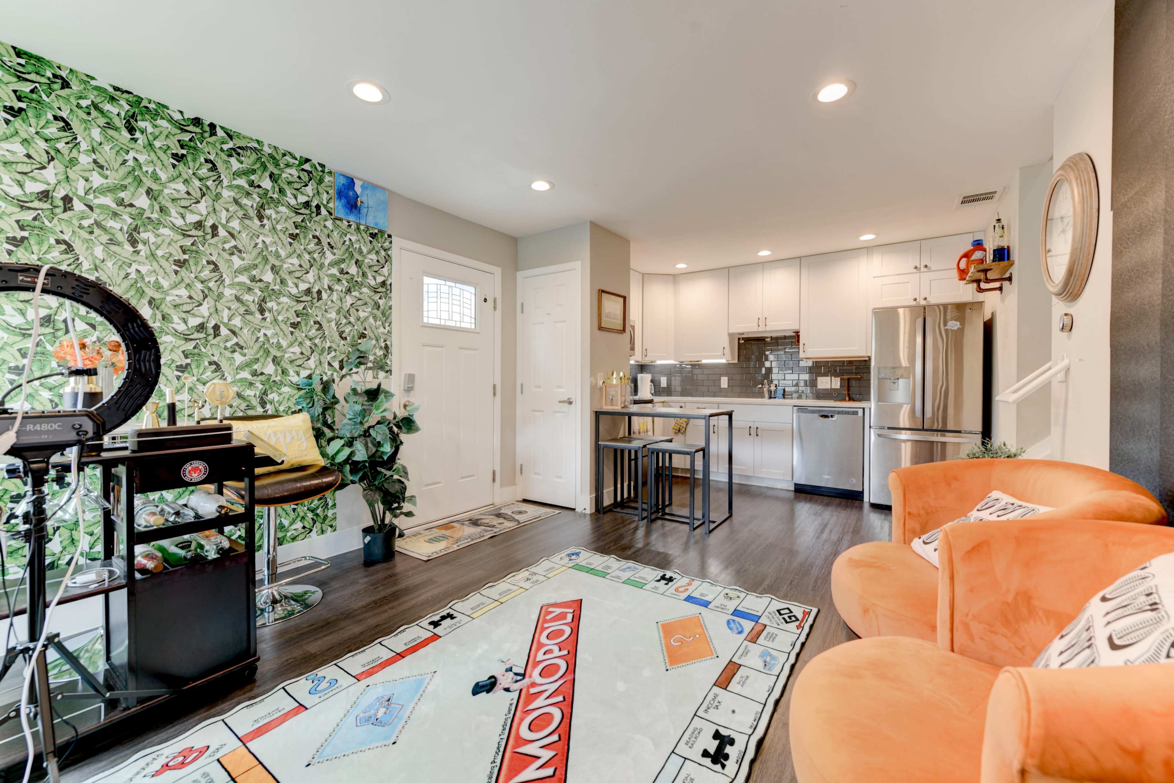 The image shows a modern living space featuring a kitchen area with a table, orange chairs, and a large Monopoly-themed rug on the floor.