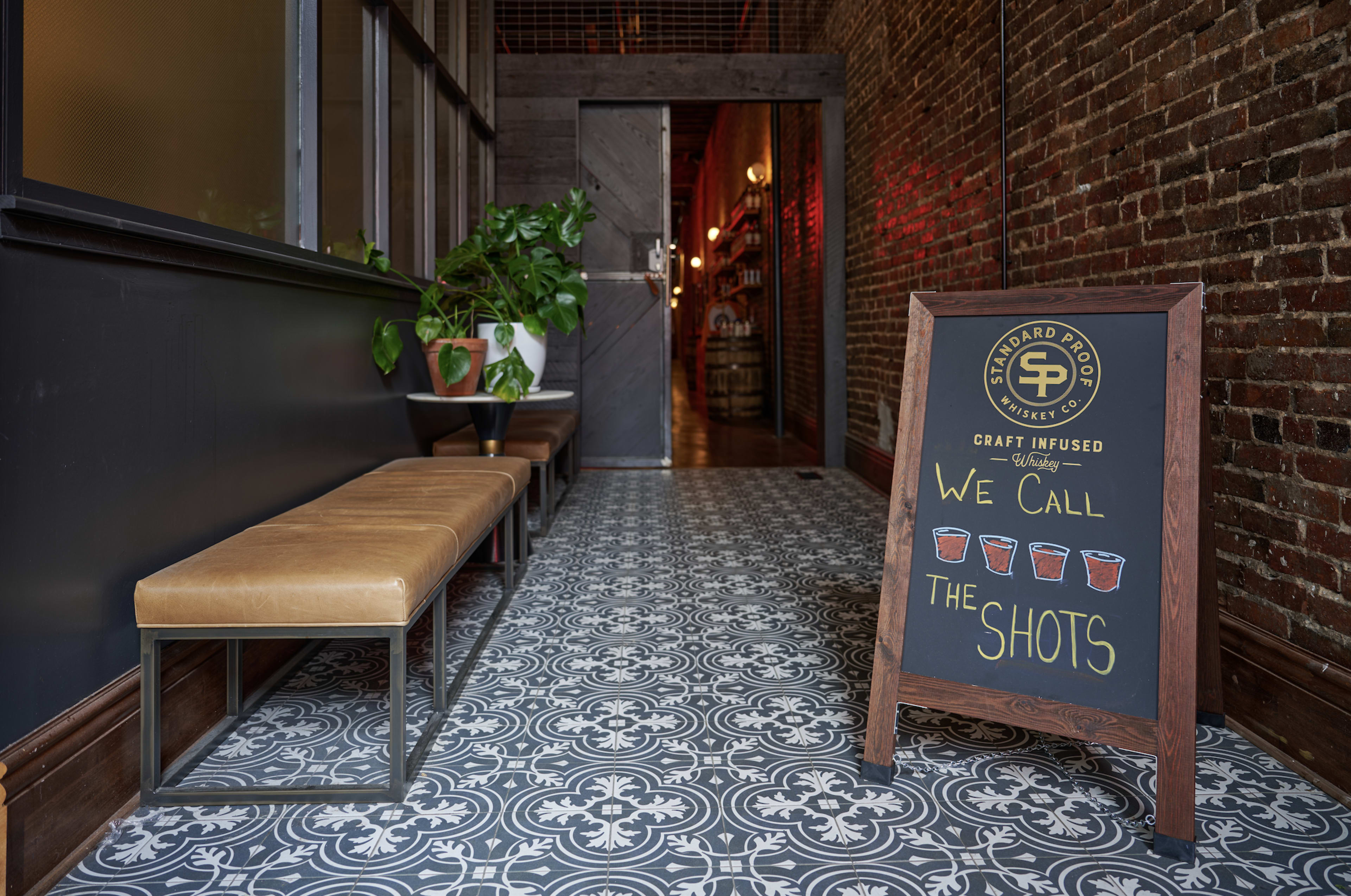The image shows a corridor with patterned tile flooring, a bench along one side, and a chalkboard sign that reads "We Call The Shots" in front of a doorway leading to another room.