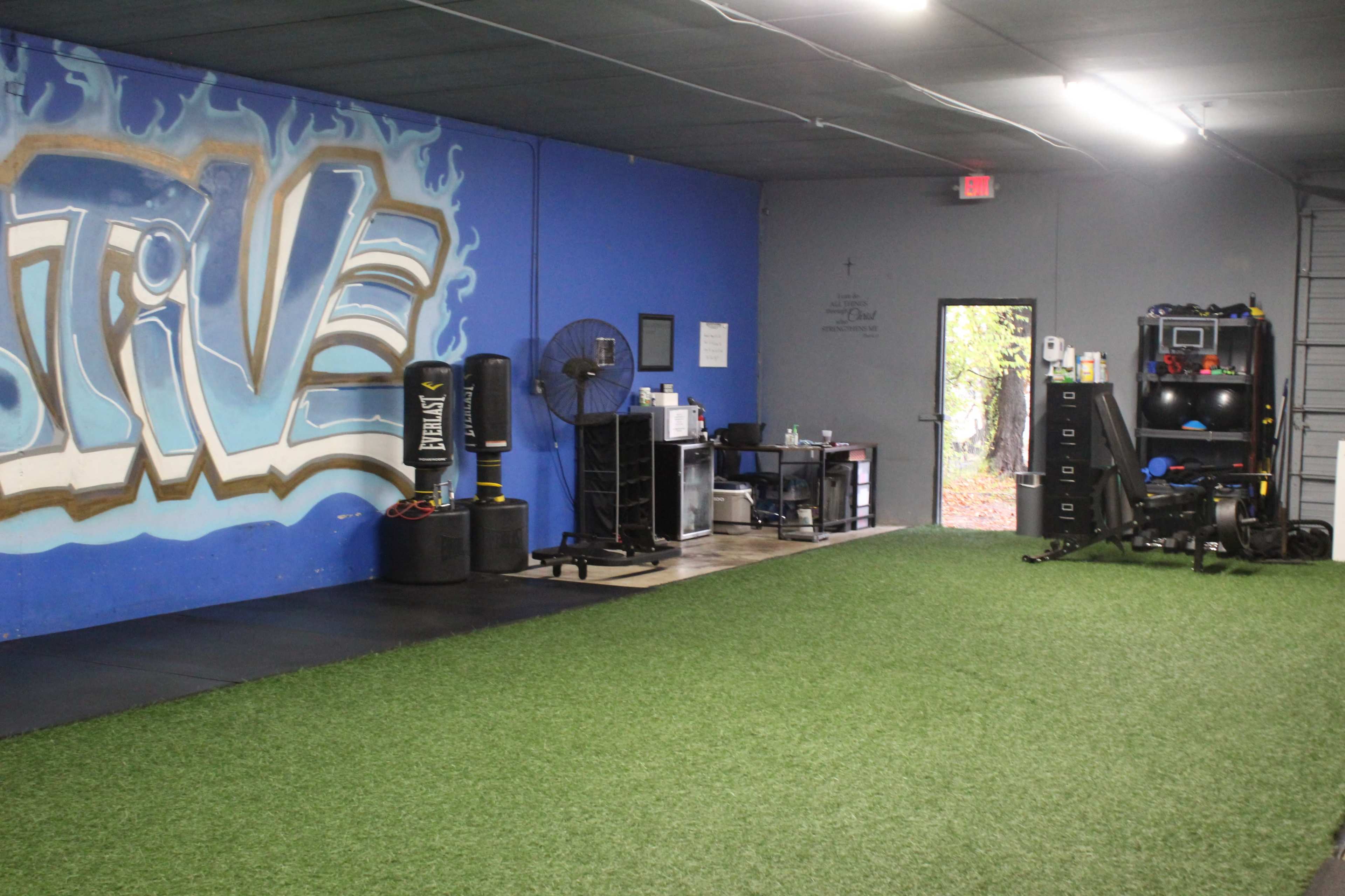 A gym interior with blue walls featuring graffiti, artificial turf flooring, and various fitness equipment along the right side.