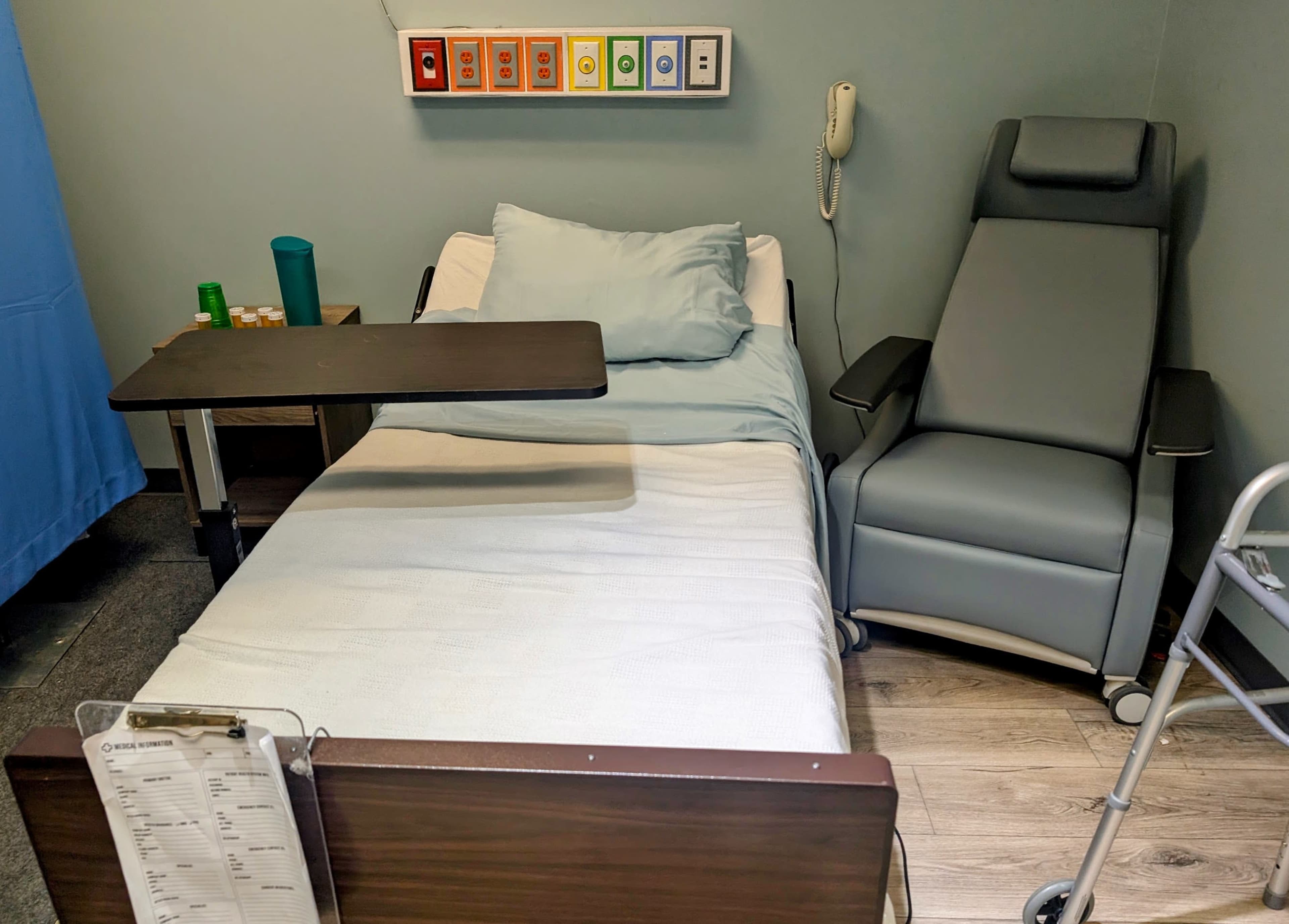 The image shows a hospital room featuring a bed with a light blue sheet, a medical reclining chair, a bedside table, and a phone mounted on the wall.