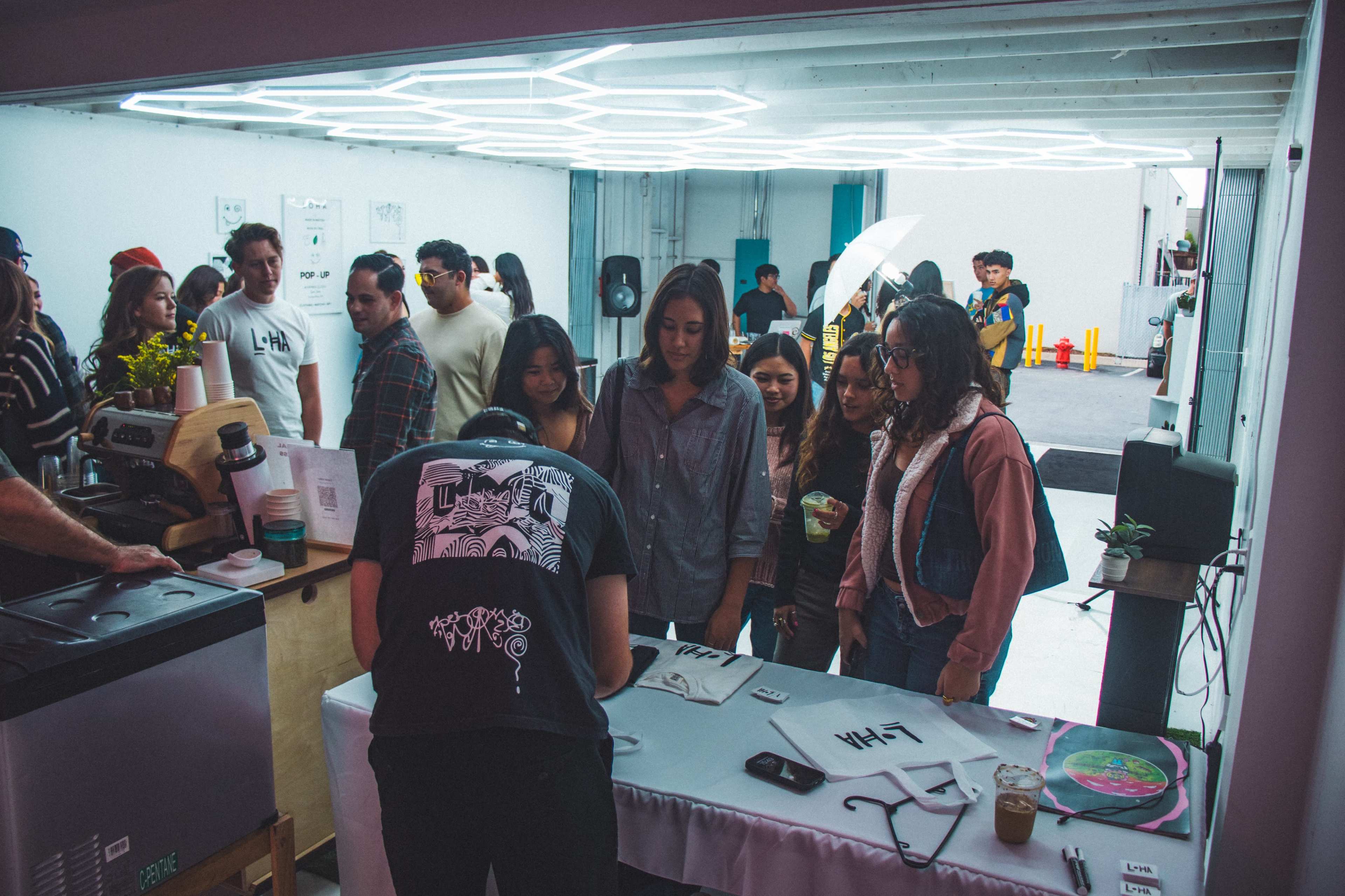 A group of people gathers around a counter in a brightly lit space, observing someone writing on a piece of paper.