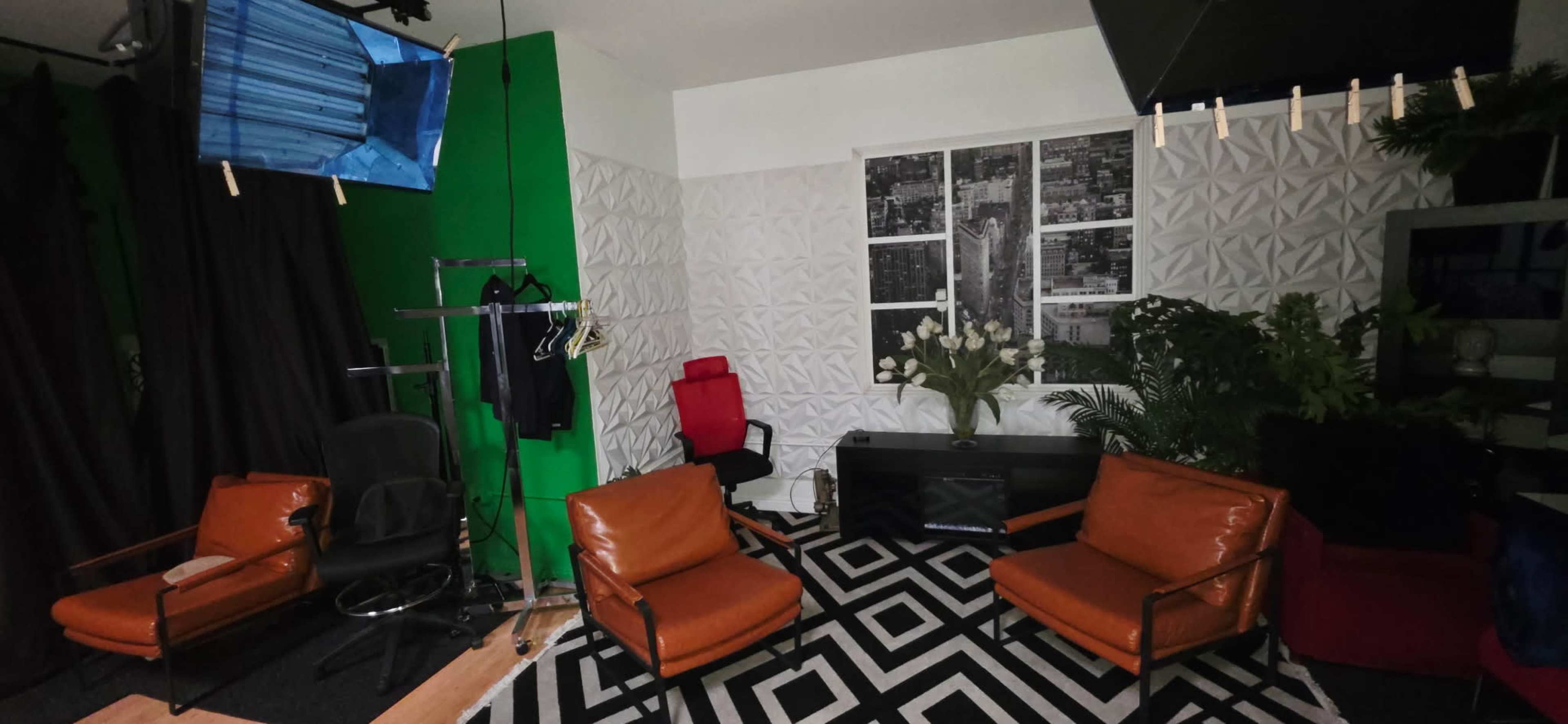 The image shows a studio setup with two orange chairs, a black and white rug, a green screen, and various equipment, including a monitor and a clothing rack.