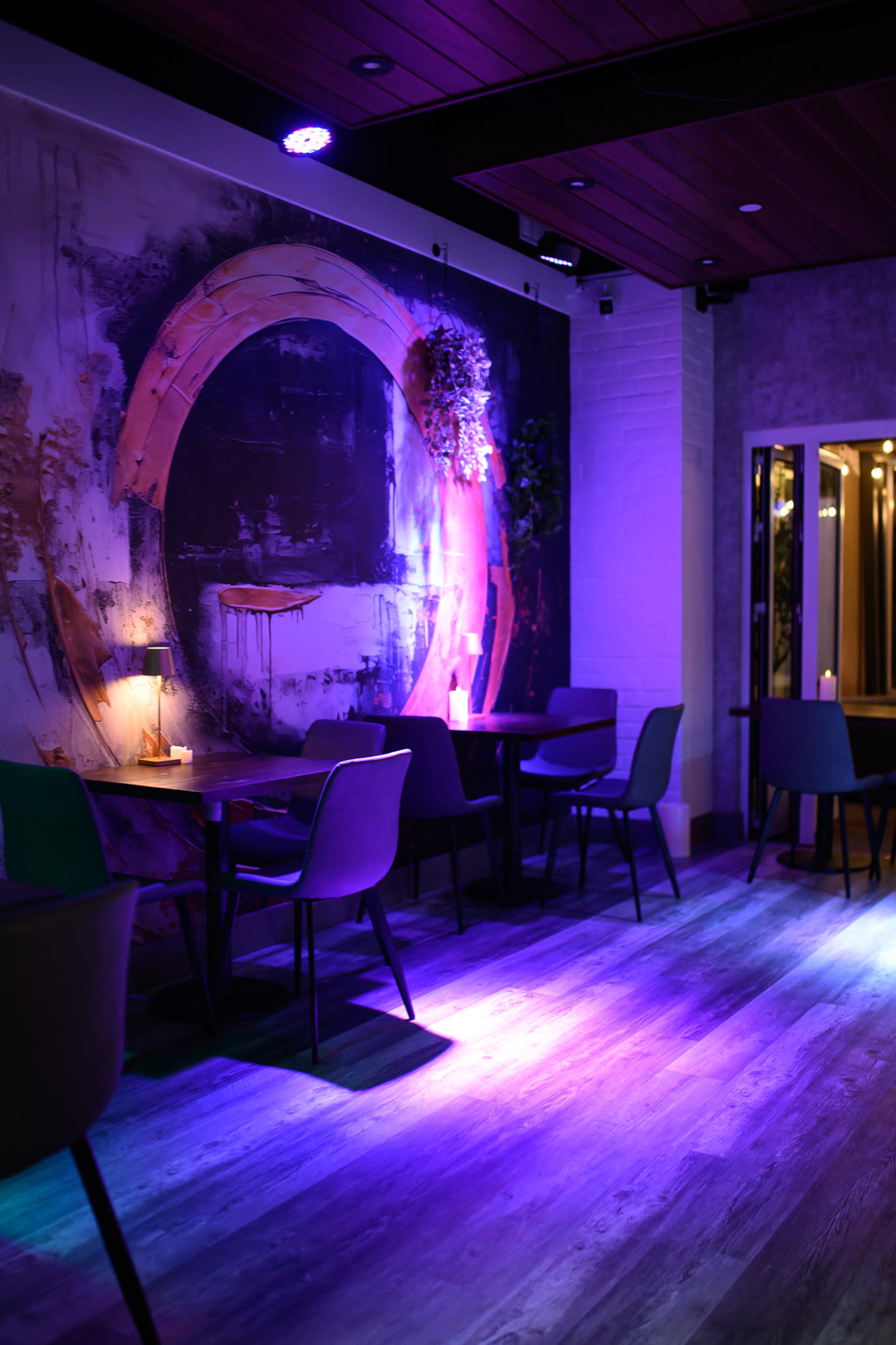The image shows an interior of a dimly lit restaurant or cafe with purple lighting, featuring modern tables and chairs against a colorful abstract mural on the wall.