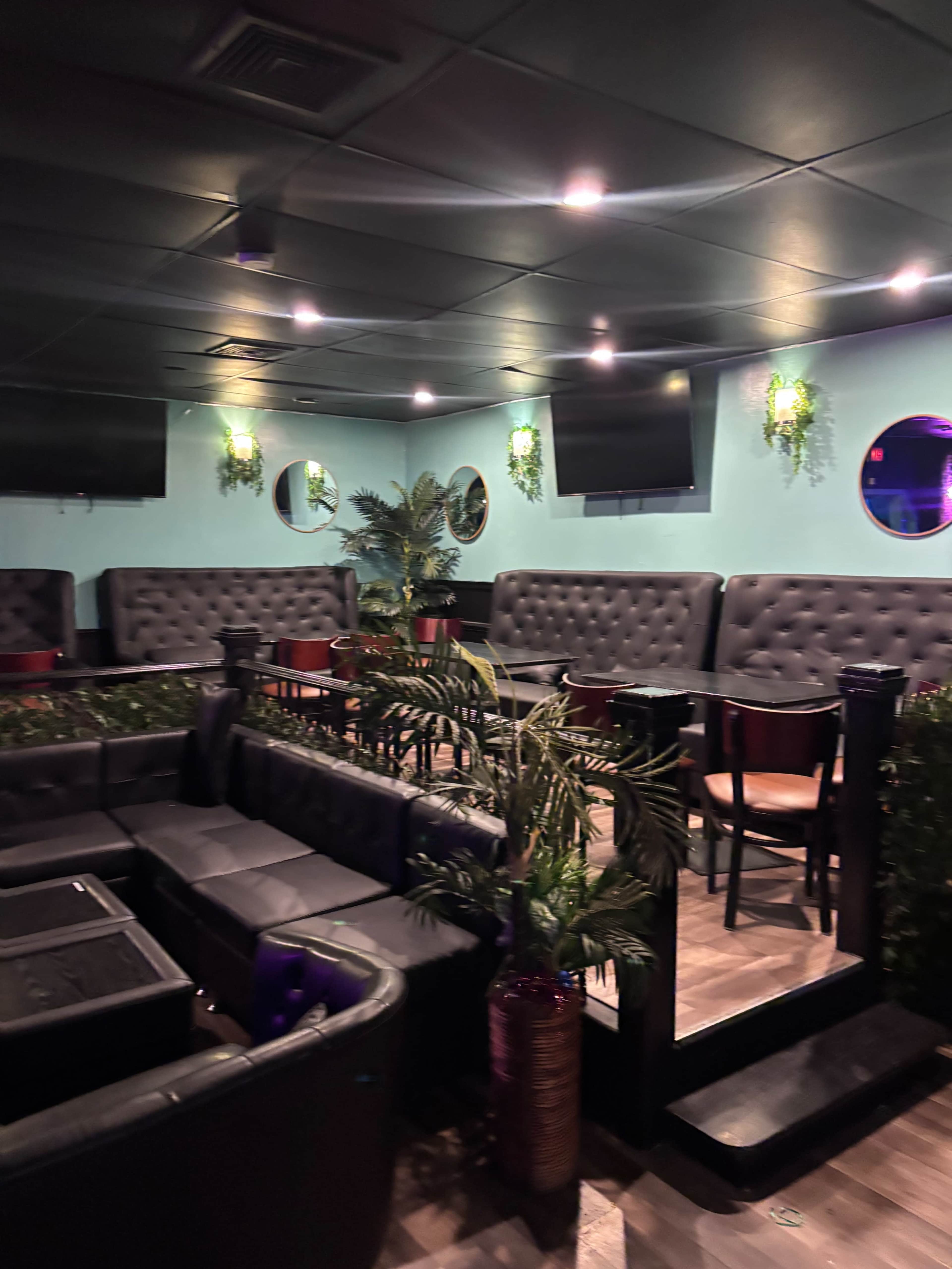 The image shows a dimly lit lounge area with black leather seating, circular mirrors on the walls, and modern decor, including potted plants.