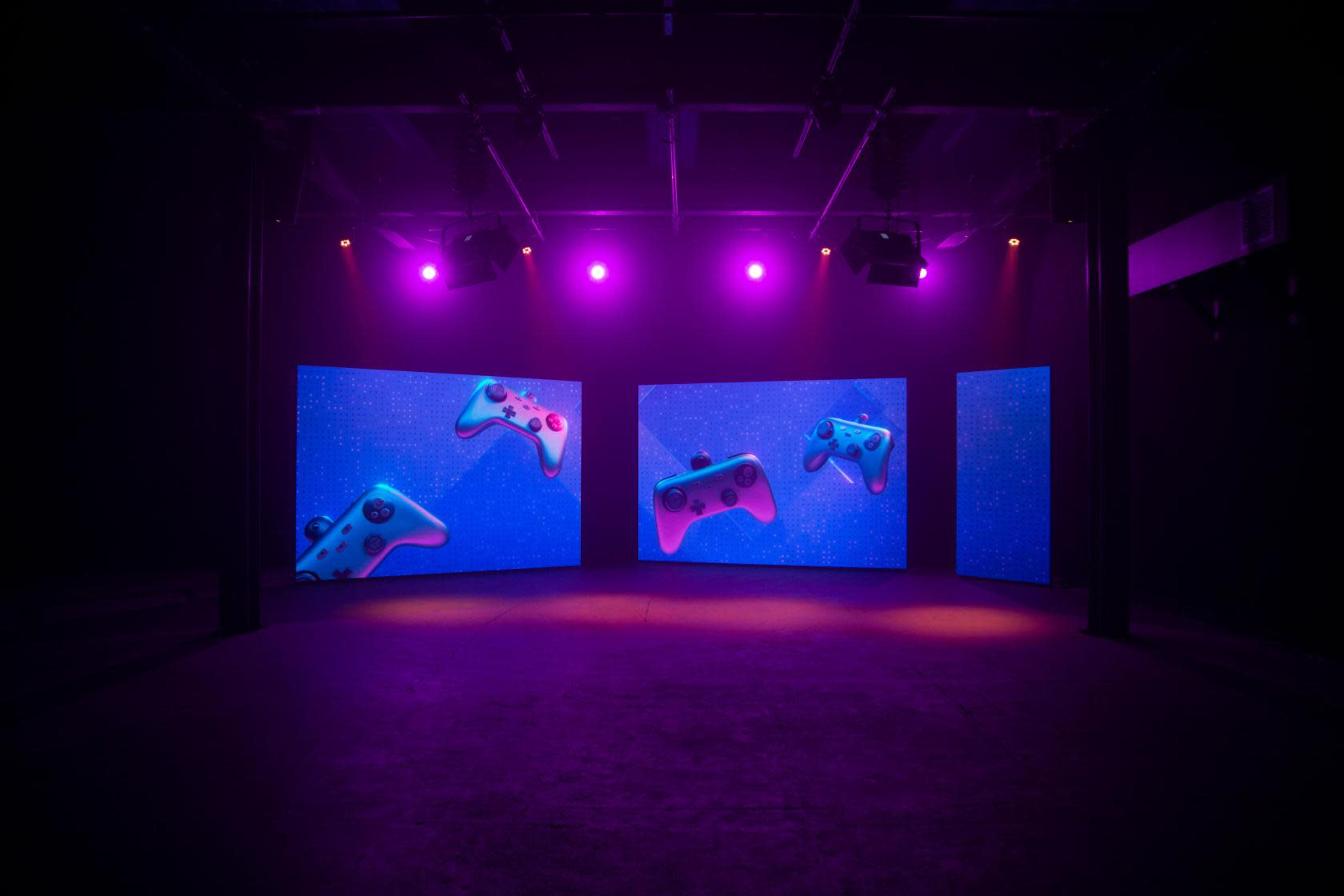 A dark room with three large screens displaying floating game controllers, illuminated by purple lights.