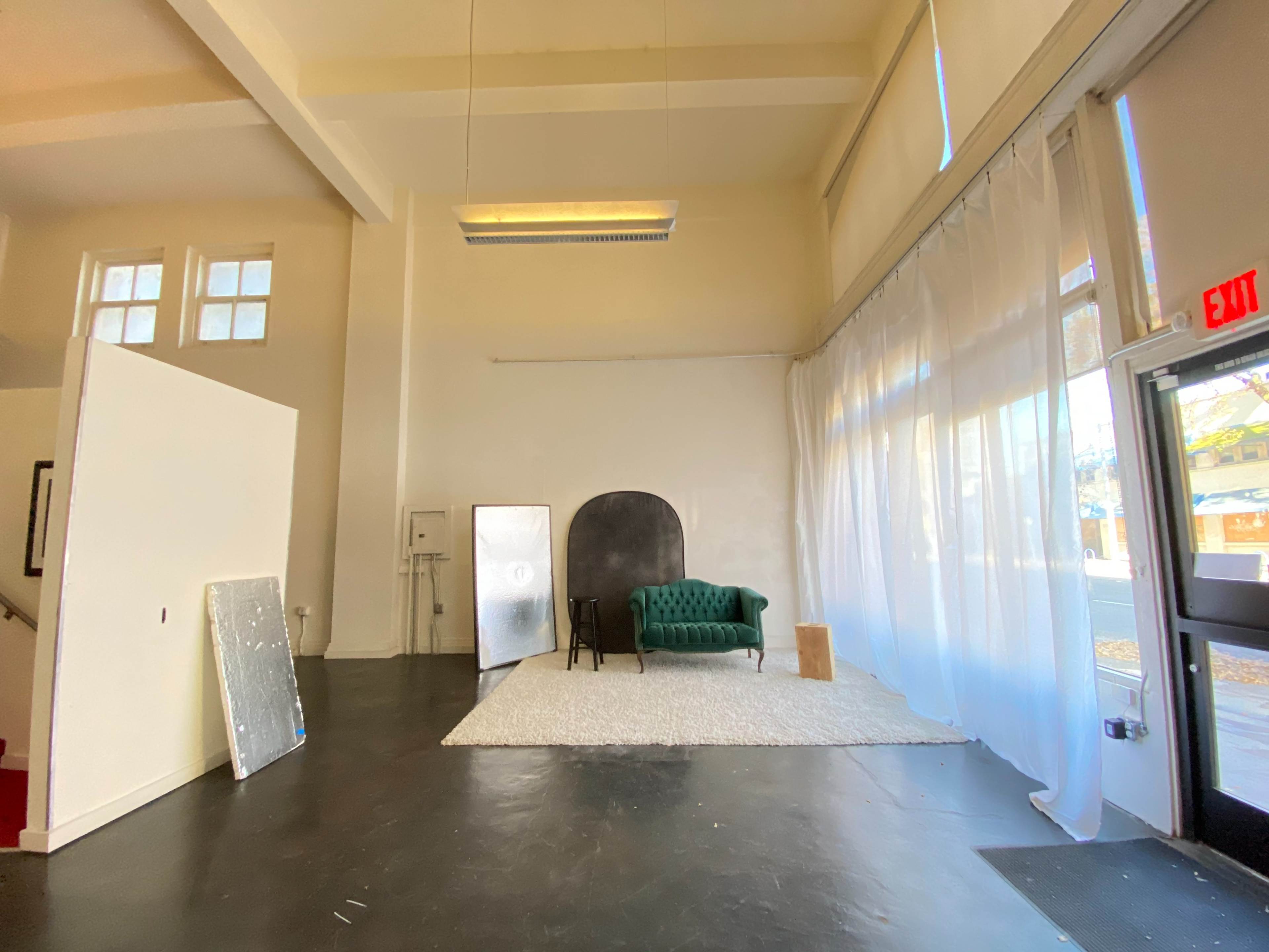 A spacious, well-lit room with a green velvet sofa, a large window covered by sheer curtains, and minimal furnishings including a rug and wall panels.