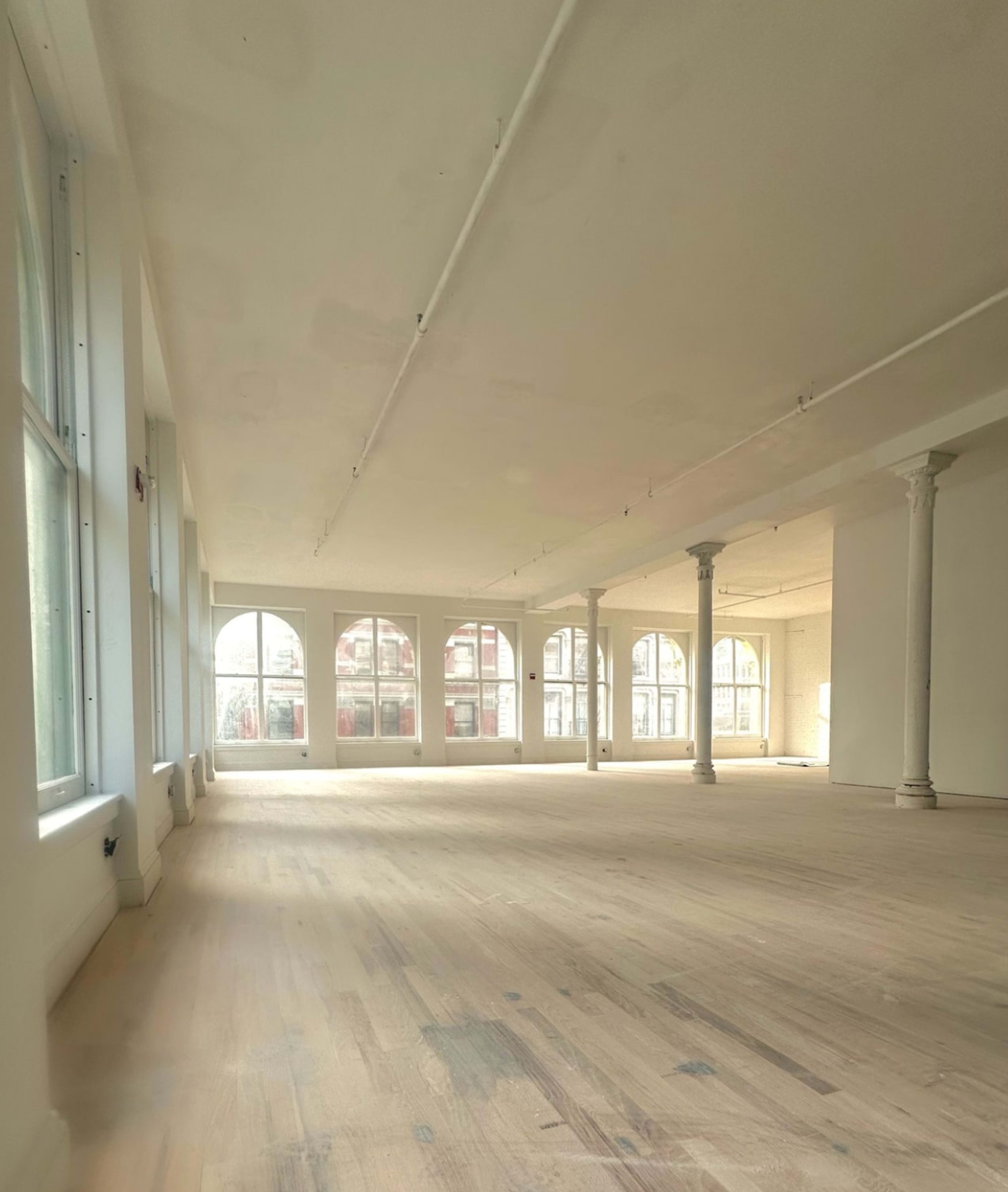 The image shows a spacious, empty interior of a room with large windows and exposed columns.