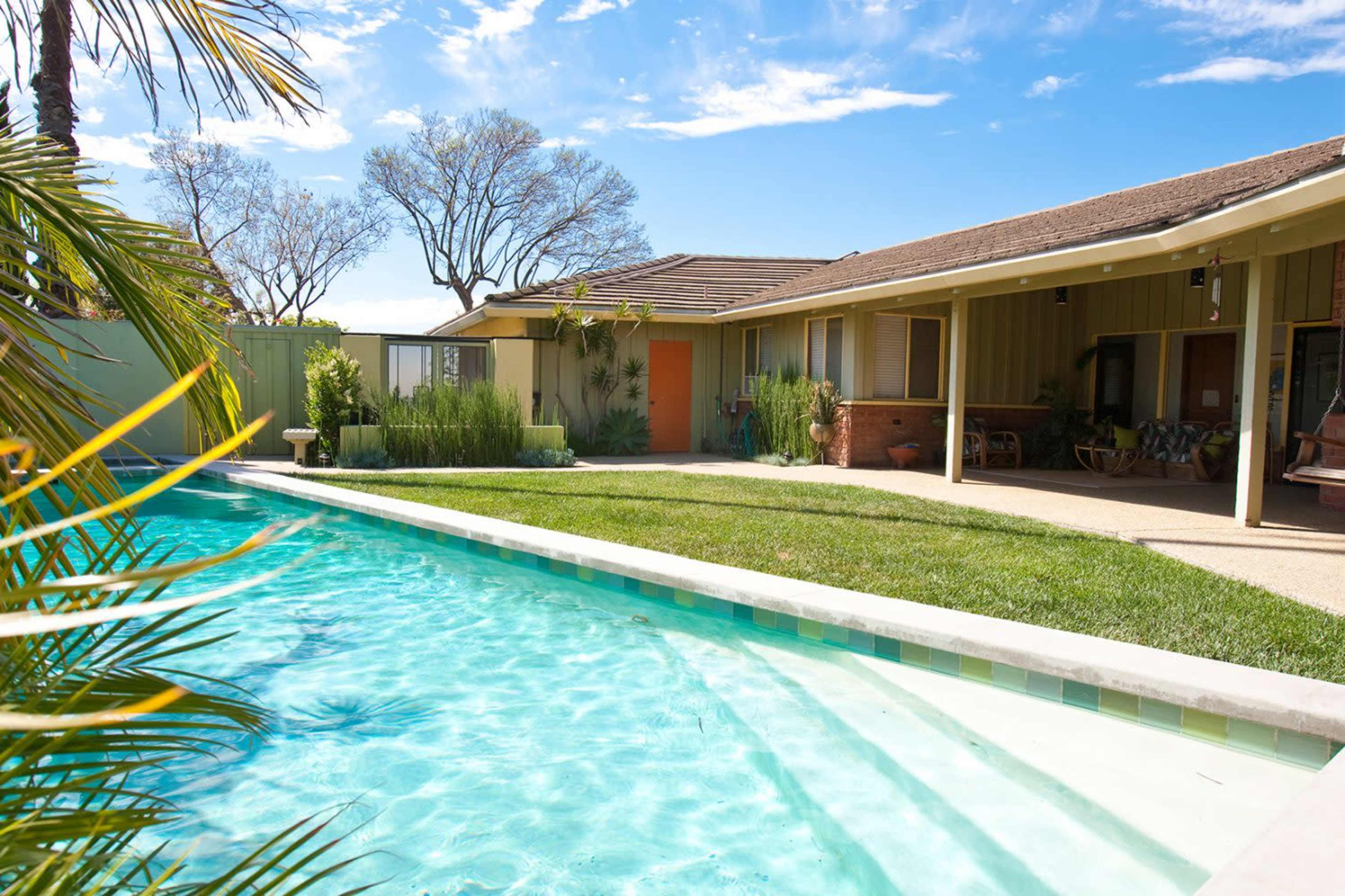 The image shows a backyard with a swimming pool, lush green grass, and a single-story house with a patio and palm trees.