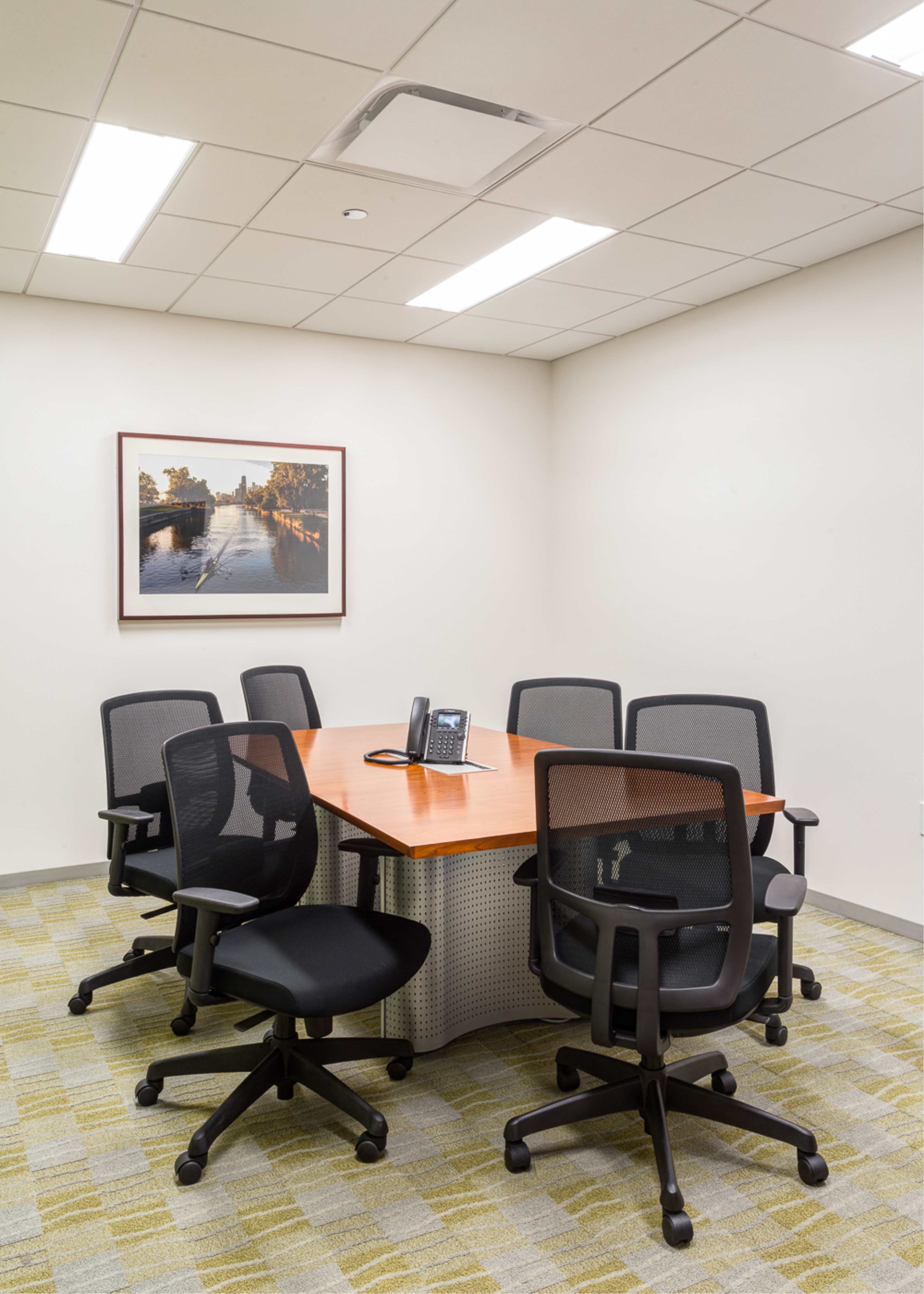 A small conference room with a wooden table surrounded by six black office chairs, a telephone, and a framed photograph on the wall.