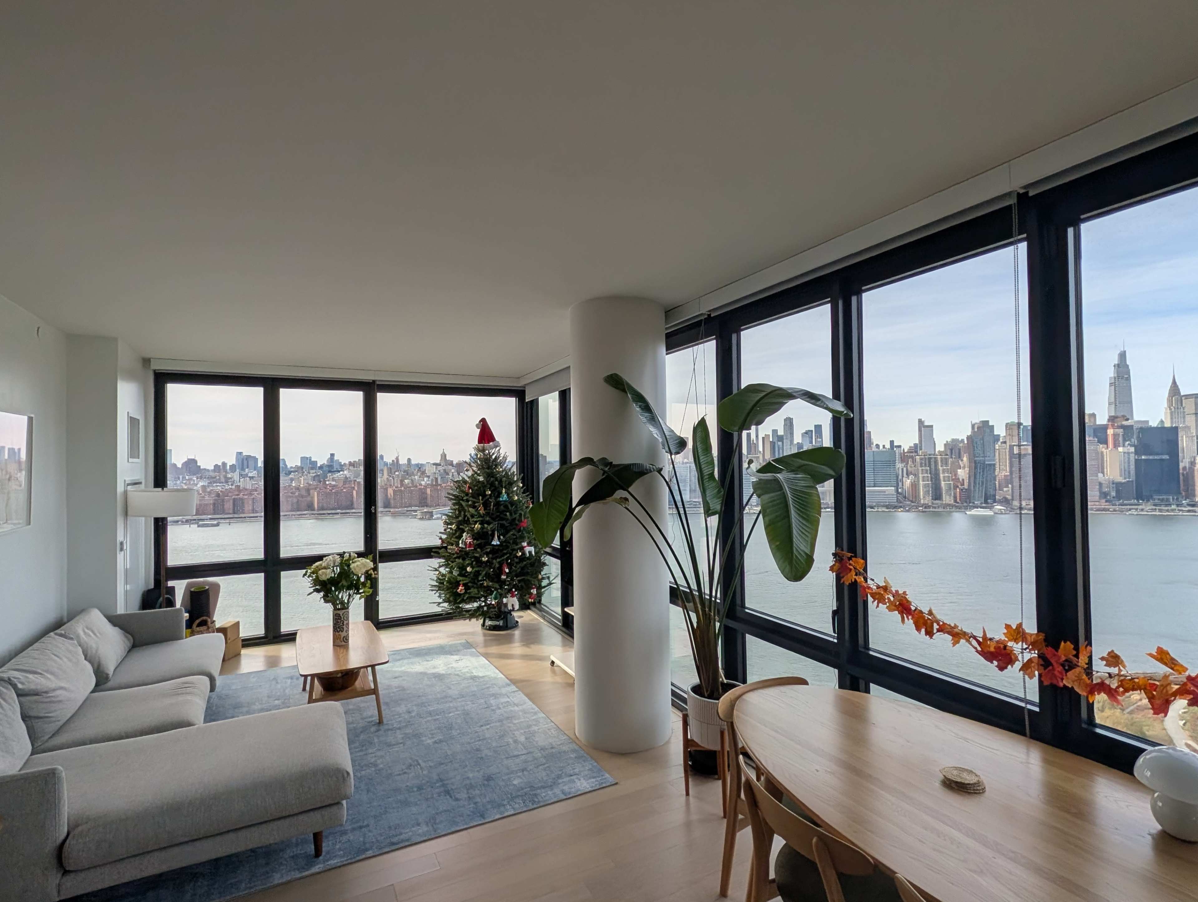 The image shows a modern living room with large windows overlooking a waterfront city skyline, a decorated Christmas tree, and a dining area with a wooden table.