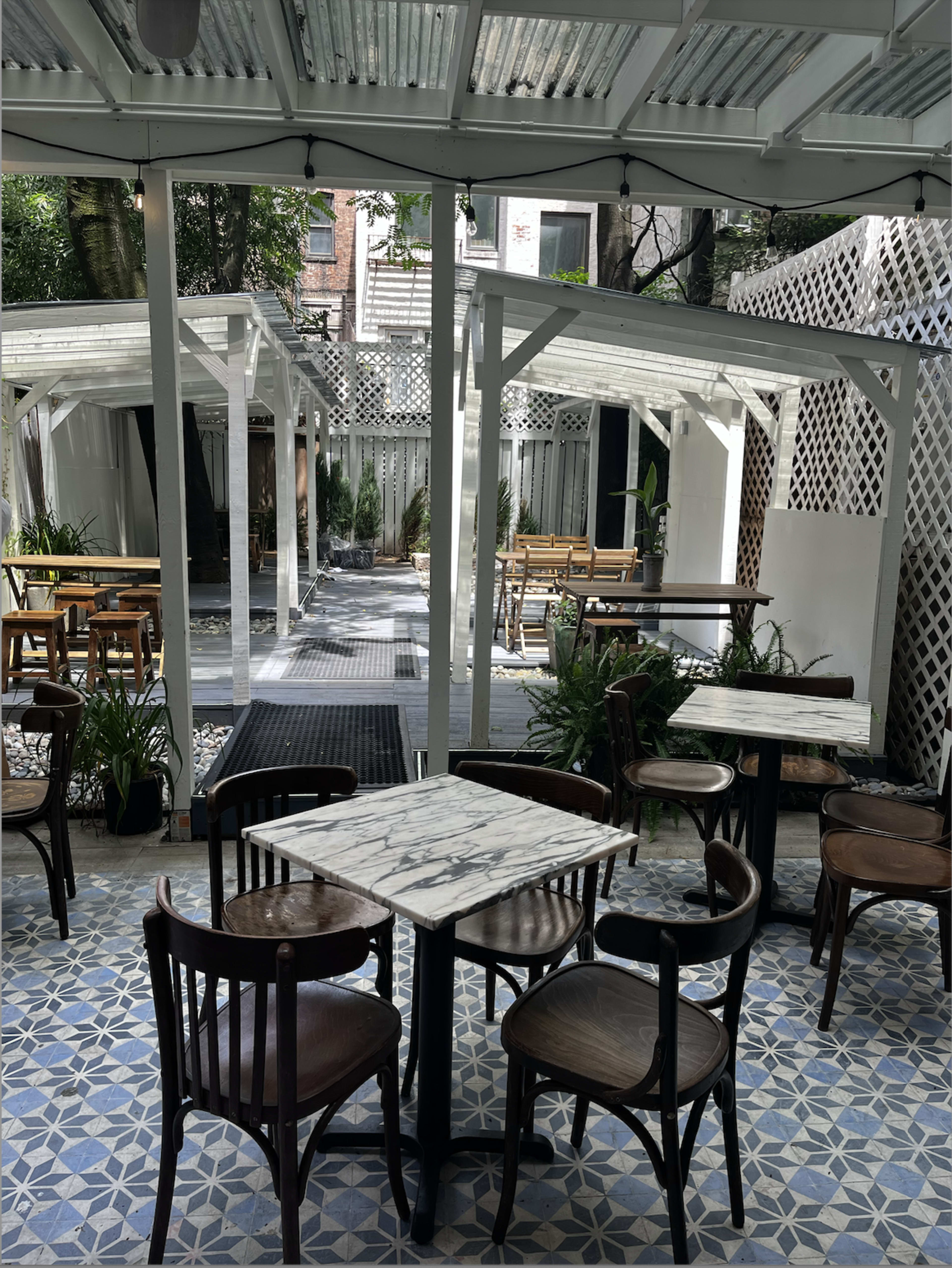 The image shows a patio area of a café with wooden tables and chairs, surrounded by greenery and an open, lattice-covered structure.