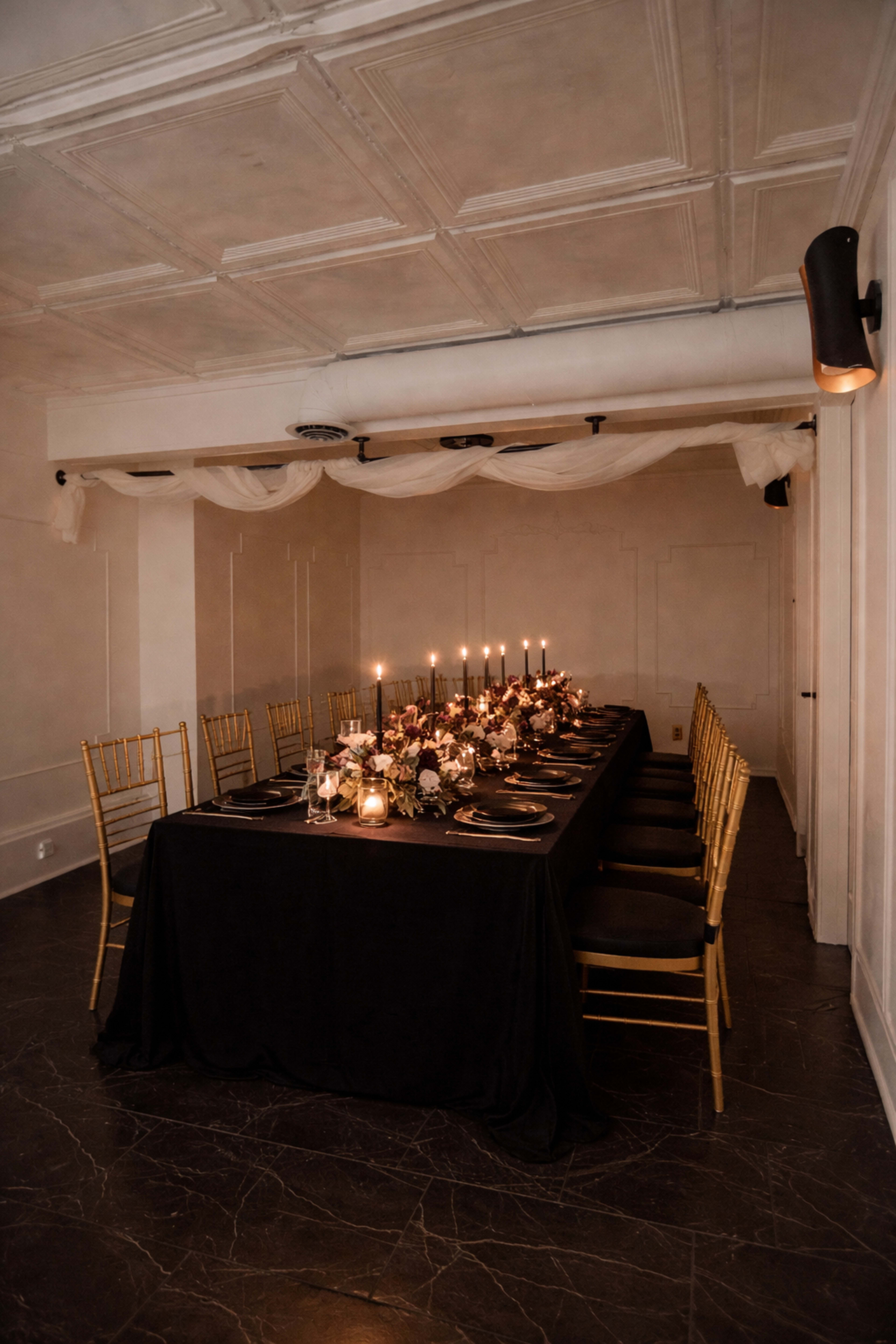 A long dining table is set with black linens, elegant dishes, and lit candles, surrounded by gold chairs in a softly lit room.