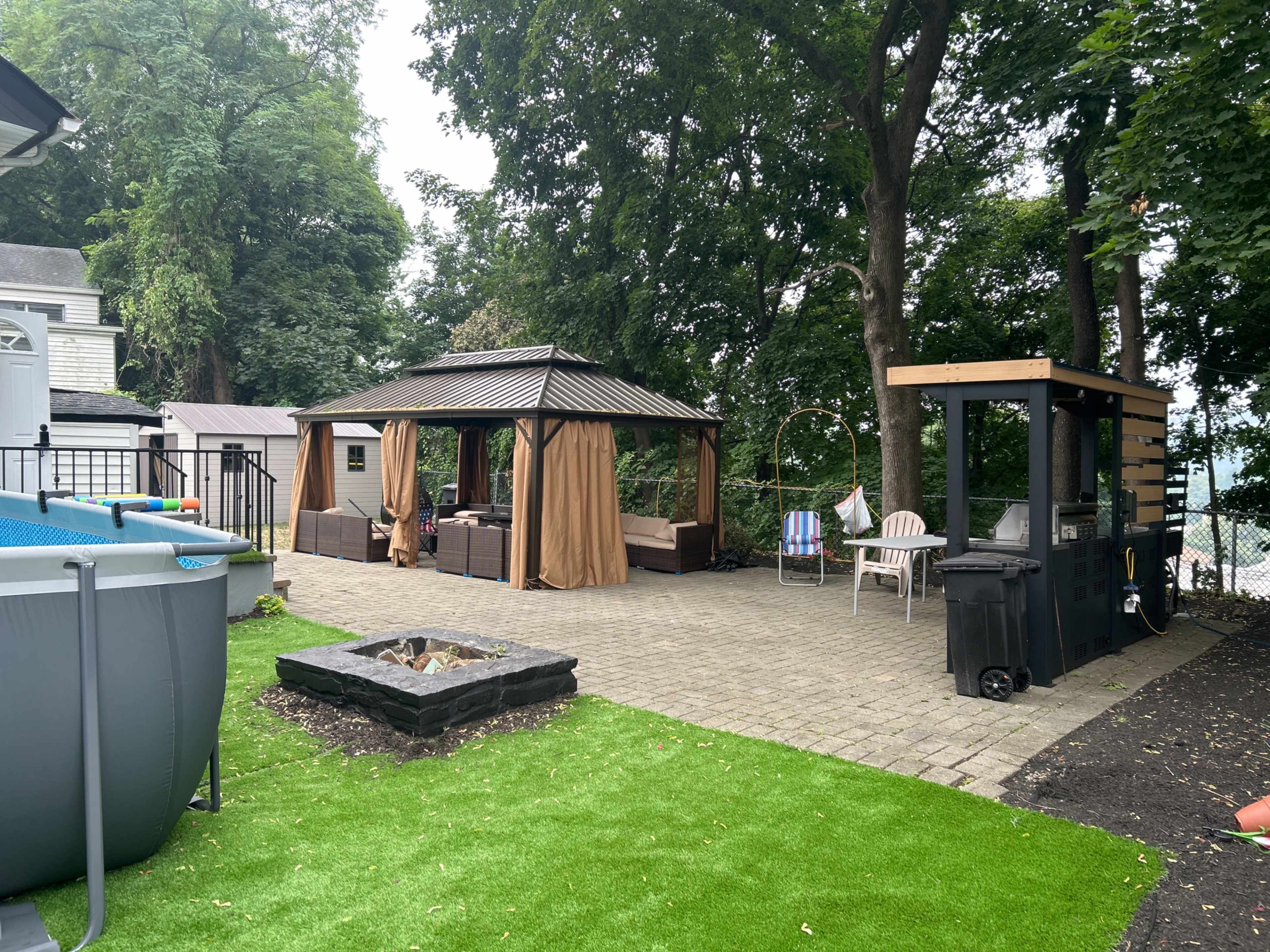 The image shows a backyard area with a gazebo surrounded by seating, a fire pit, and a pool, all set on a paved surface with patches of artificial grass.