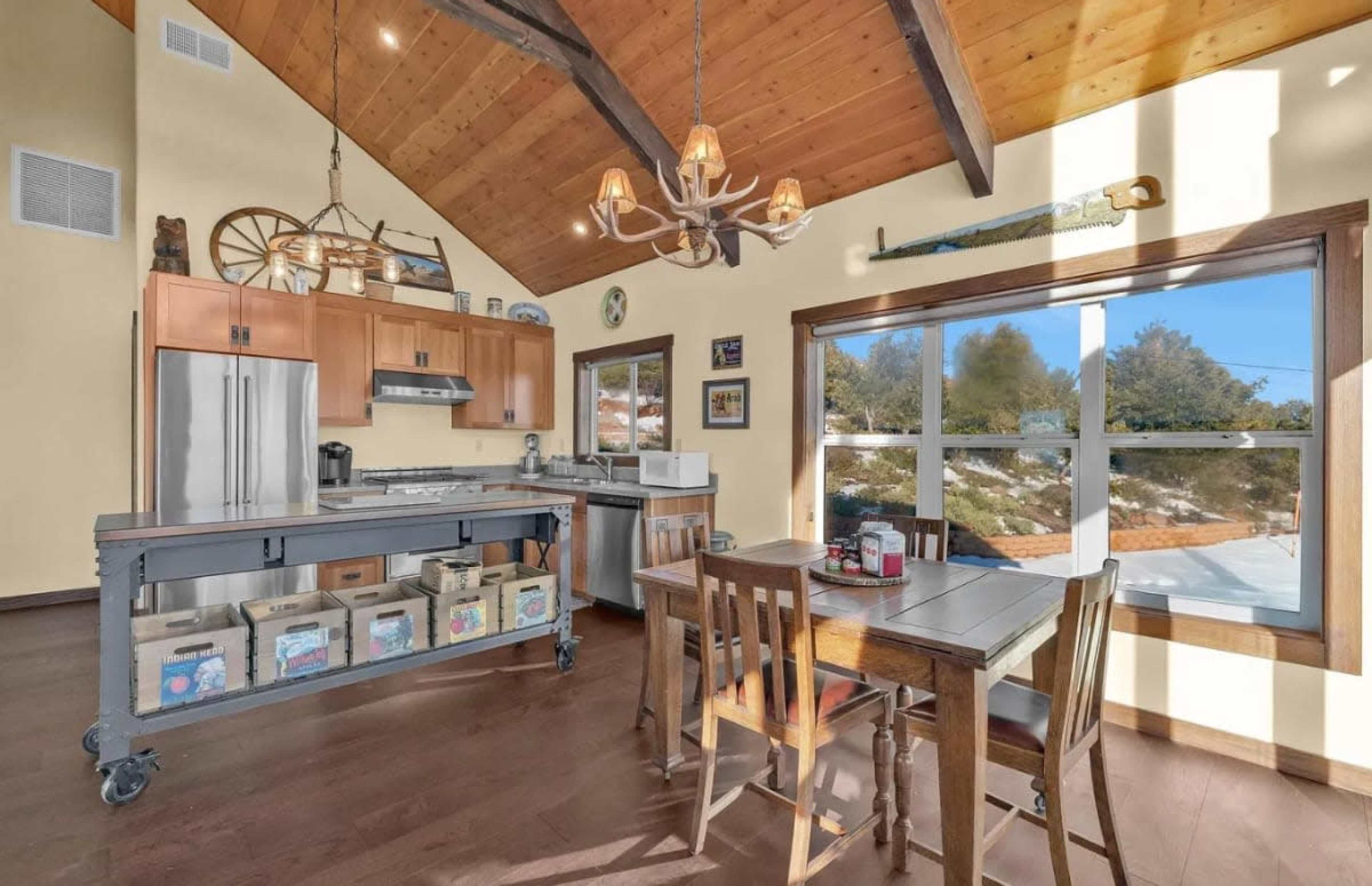 The image shows a modern kitchen with a wooden ceiling, stainless steel appliances, and a dining table set next to a large window.