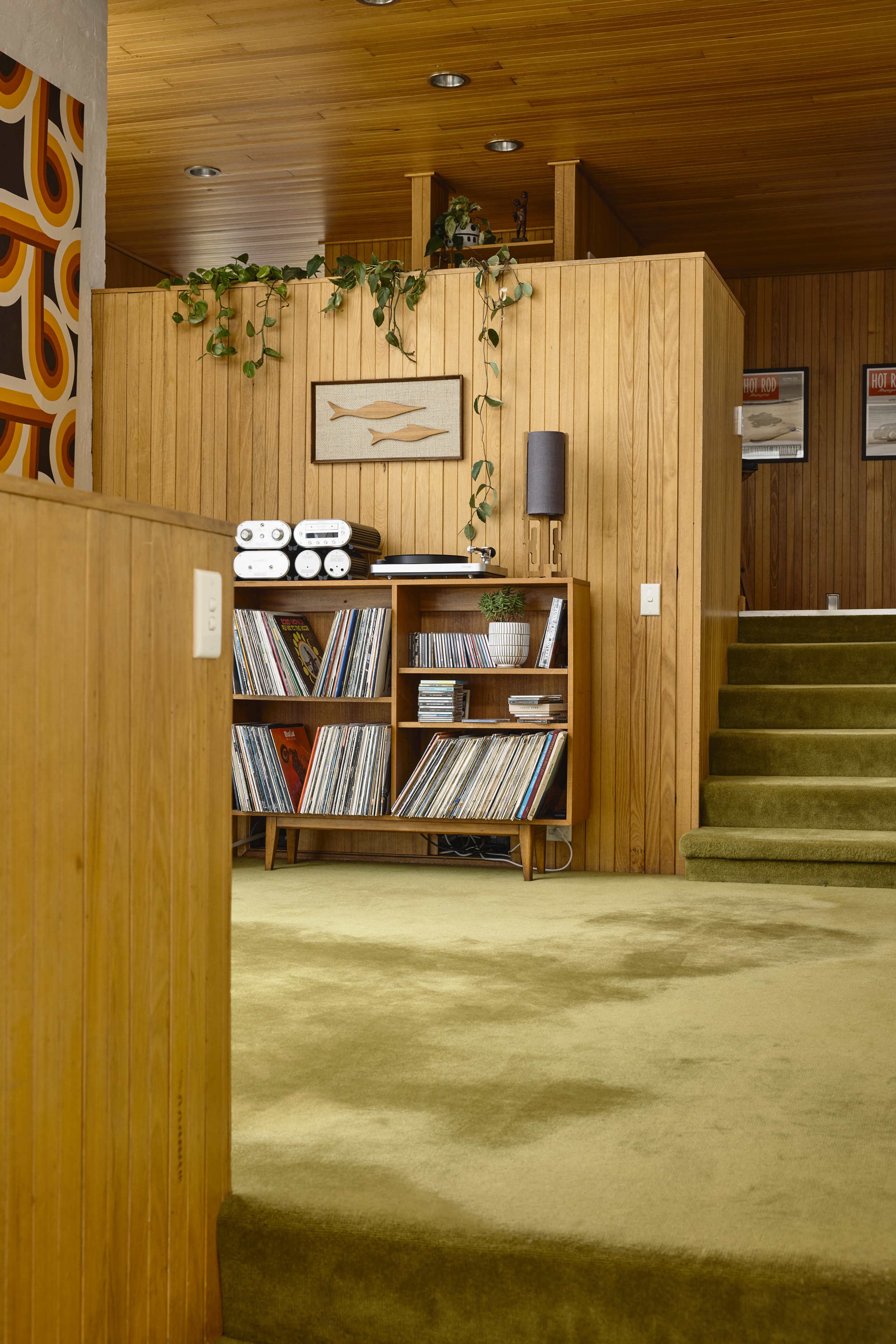 The image shows a mid-century modern interior featuring wooden paneling, a record shelf filled with vinyl records, and green carpeting leading up a set of stairs.