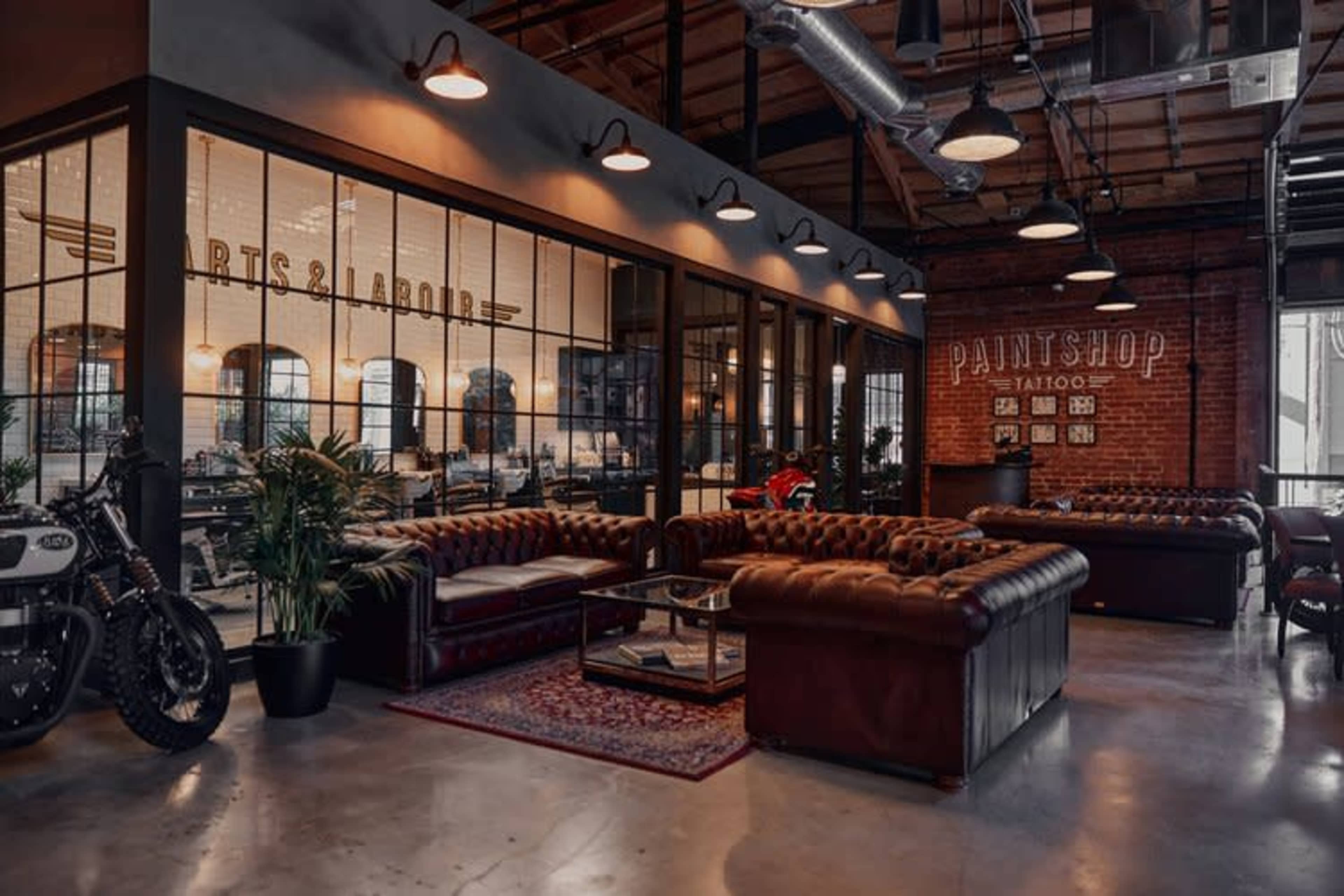 A modern tattoo studio featuring leather couches, industrial decor, and large glass windows that provide a view of the workspace.