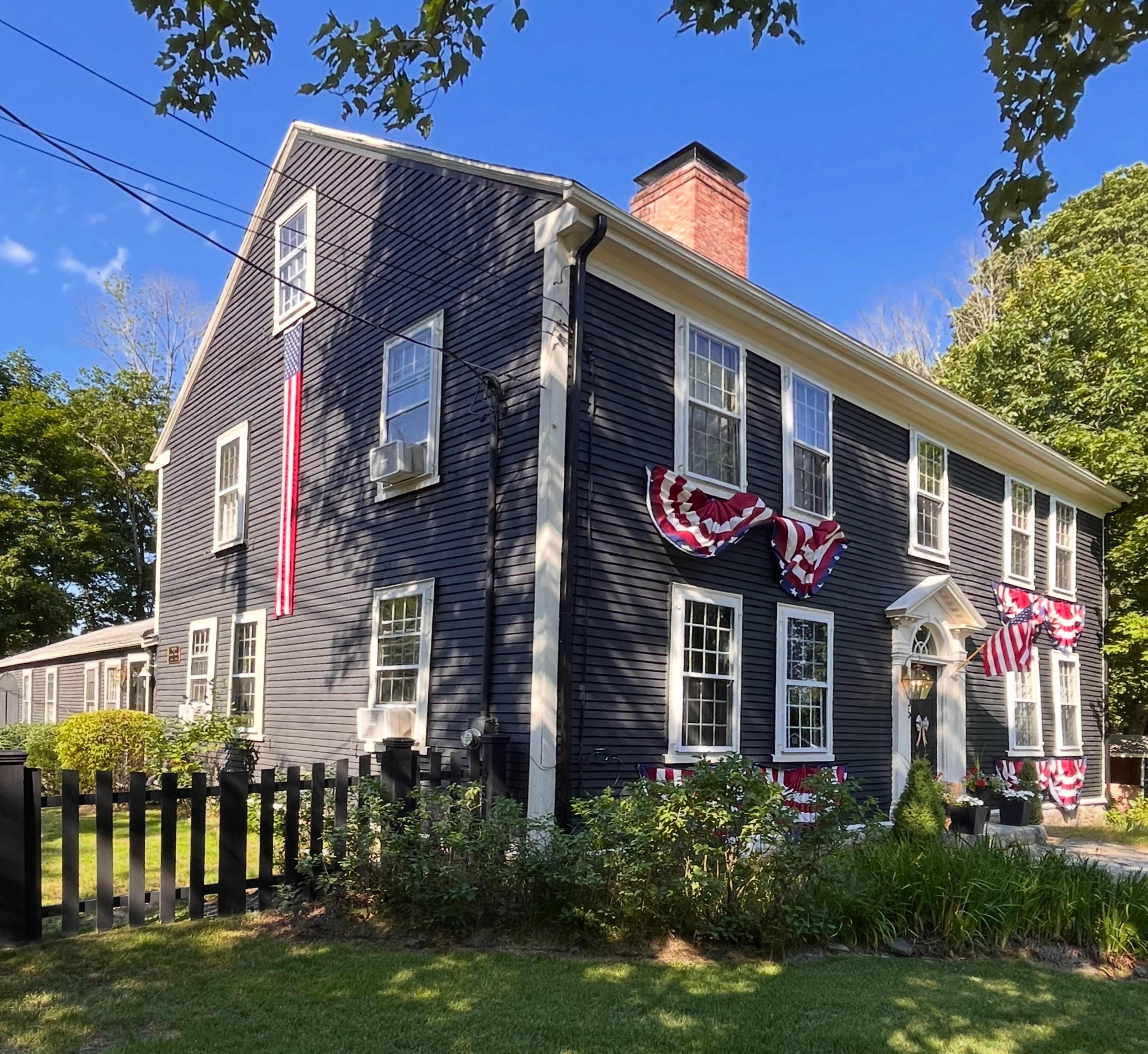 A two-story blue house with white trim is adorned with red, white, and blue bunting, surrounded by a well-maintained lawn and a black picket fence.