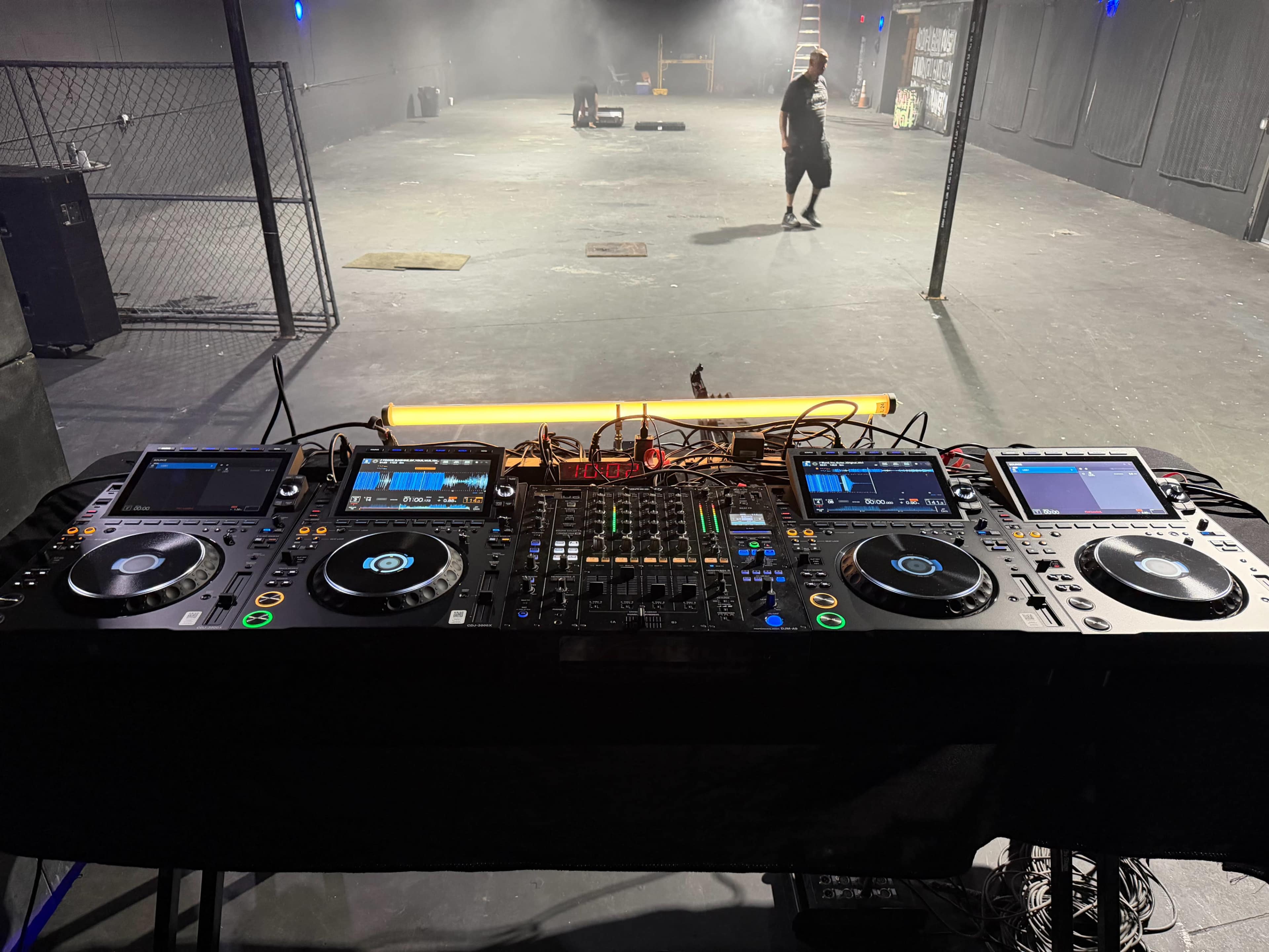 A DJ setup featuring two turntables and a mixer is positioned in the foreground of an empty, dimly lit performance space.