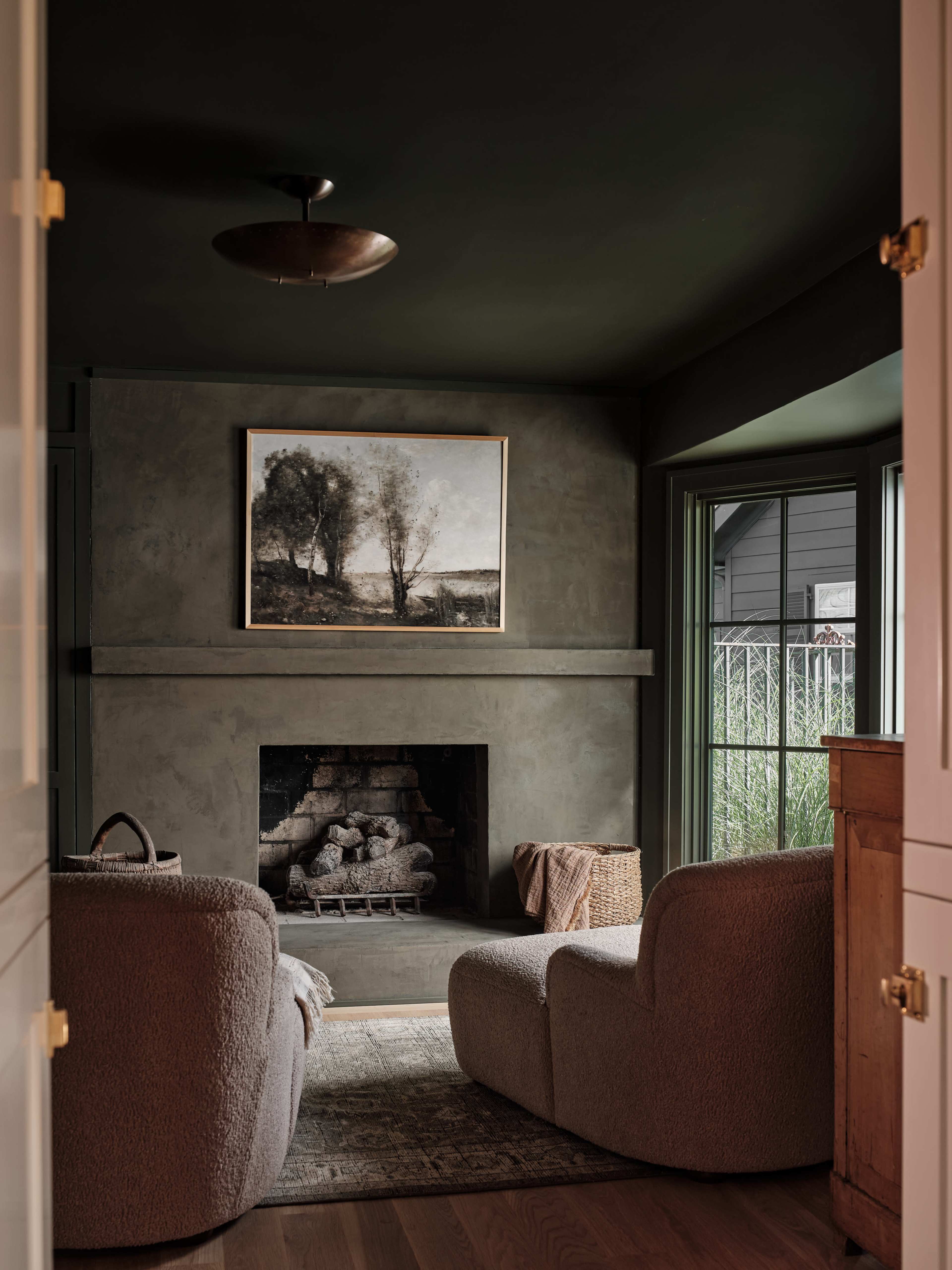 A cozy living room features two curved chairs facing a fireplace, with a large landscape painting above and windows providing natural light.