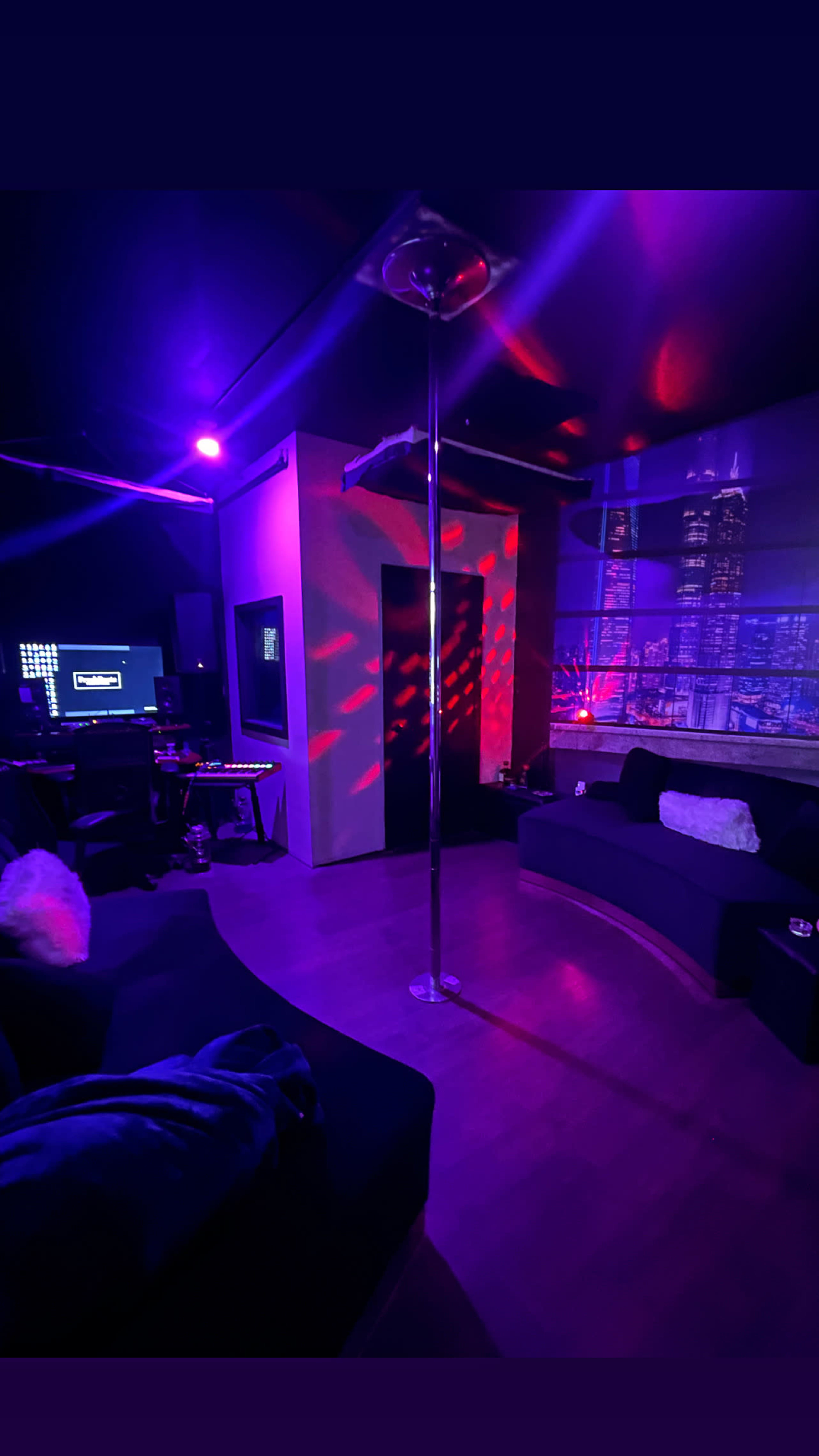 The image shows a dimly lit room featuring a pole in the center, surrounded by a seating area and illuminated by colorful lights projected on the walls.