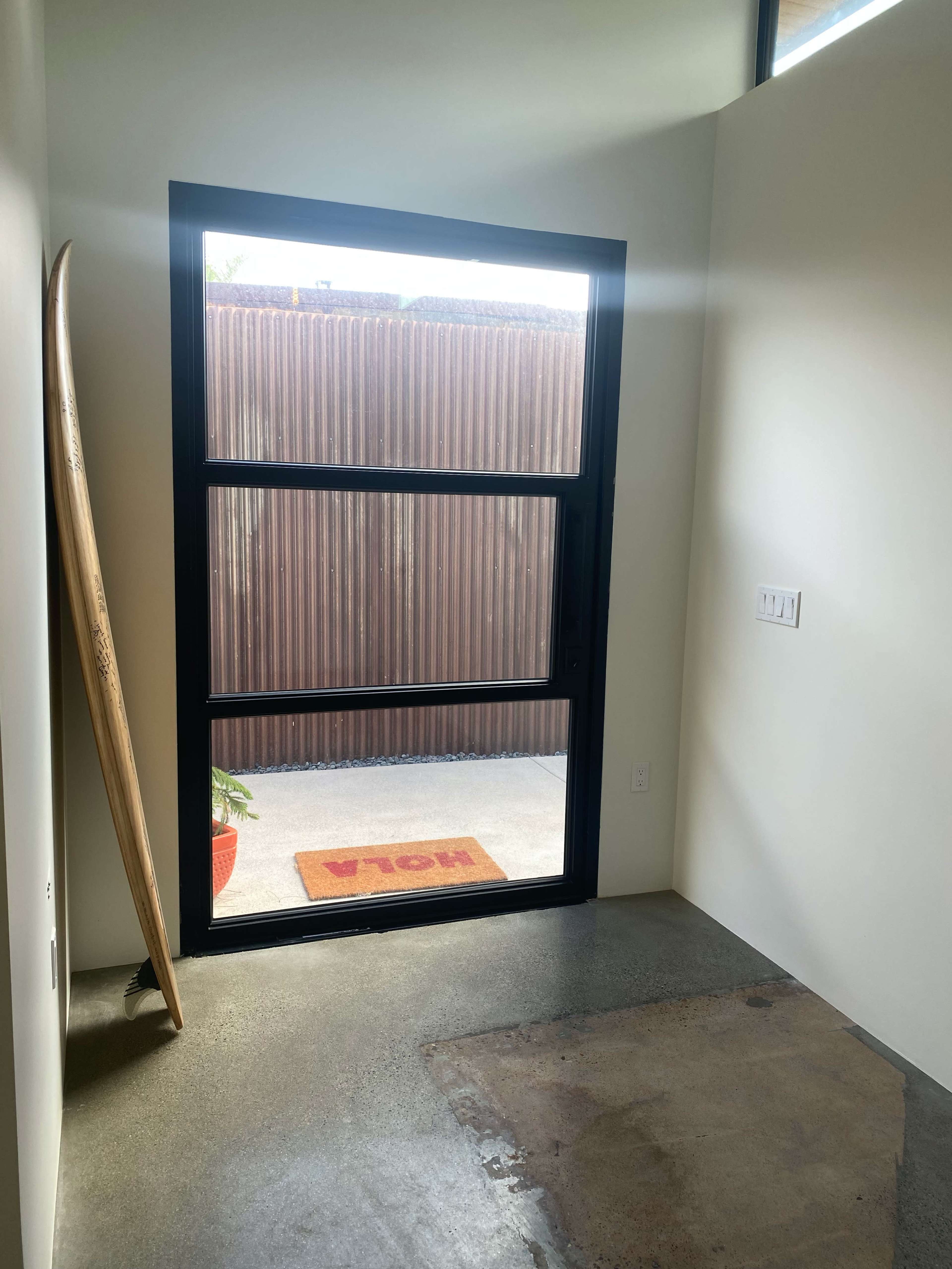 A corner of a minimalist room with a large black-framed window, a surfboard leaning against the wall, and a welcome mat at the entrance.